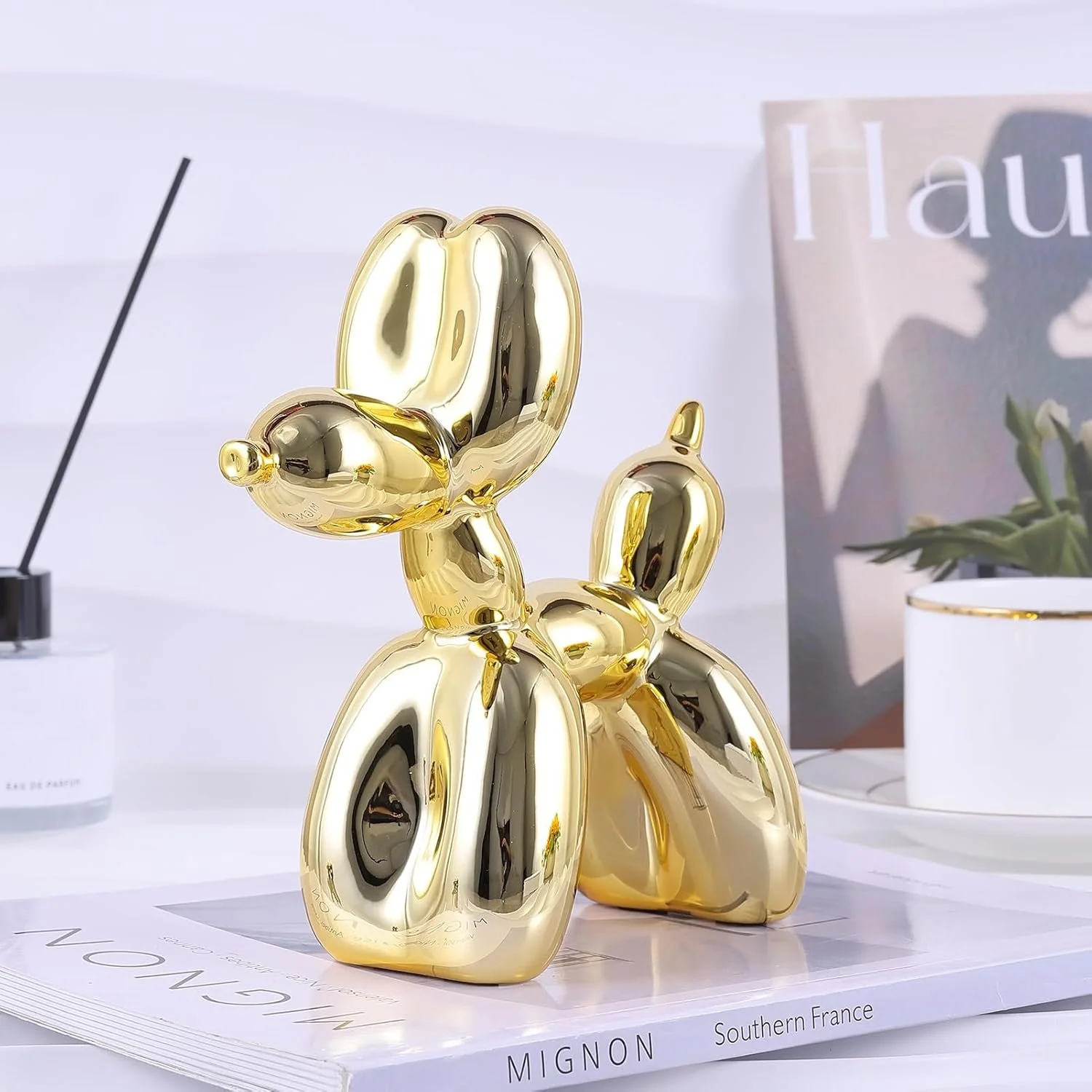 A shiny gold balloon sculpture resembling a dog, placed on a magazine on a white surface with a potted plant and a magazine in the background.