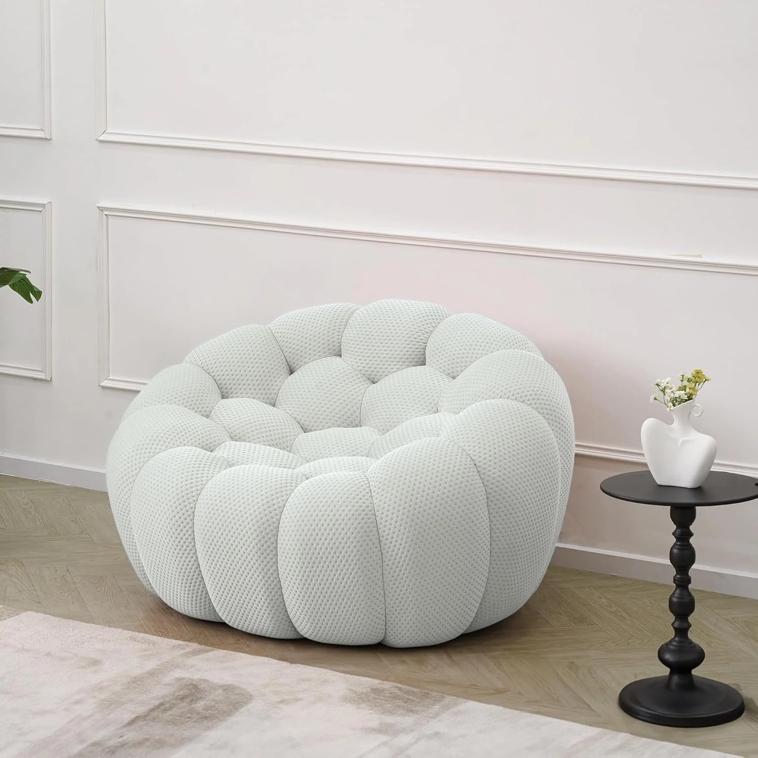 A modern white upholstered armchair with rounded, padded segments, placed next to a small black side table with a white vase holding dried flowers, in a room with white paneled walls and light wood flooring.