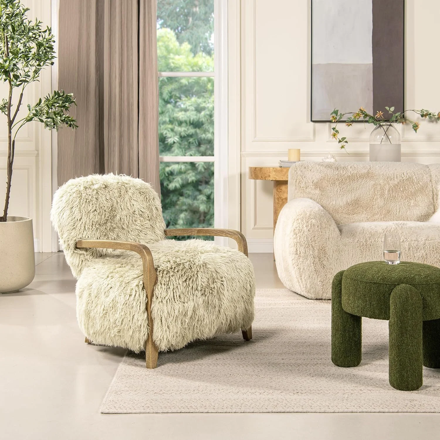 A cozy living room with a fluffy, cream-colored armchair, a white rounded sofa, and a green plush ottoman, all on a beige rug. There is a tall plant in a pot and a window with beige curtains showing green trees outside.