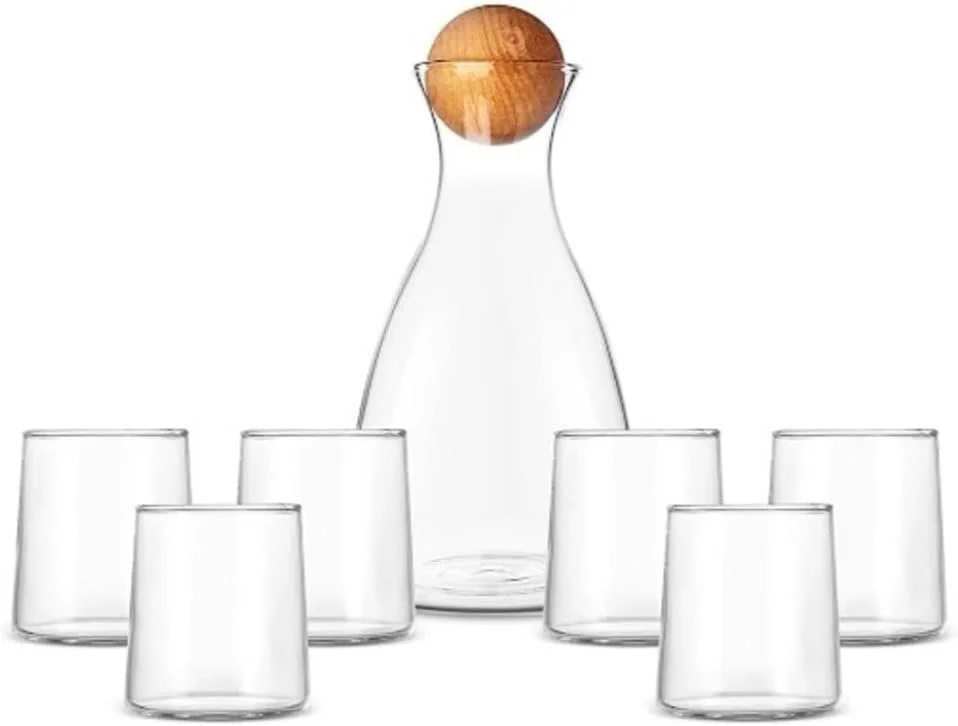 A glass carafe with a wooden stopper surrounded by six empty short glasses on a white background.