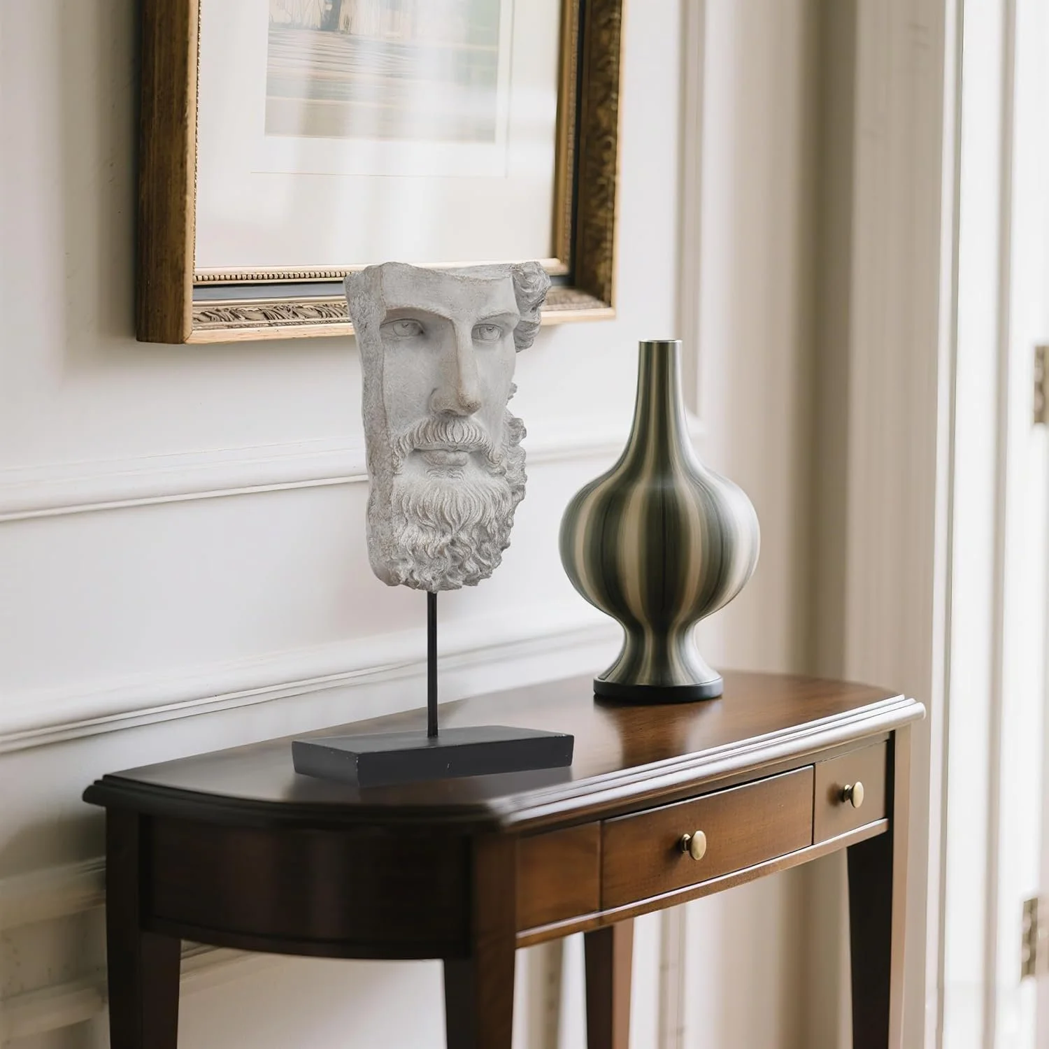 Decorative arrangement on a wooden table with a carved stone bust of a man with a beard and mustache, a striped ceramic vase, and a framed picture on the wall behind.