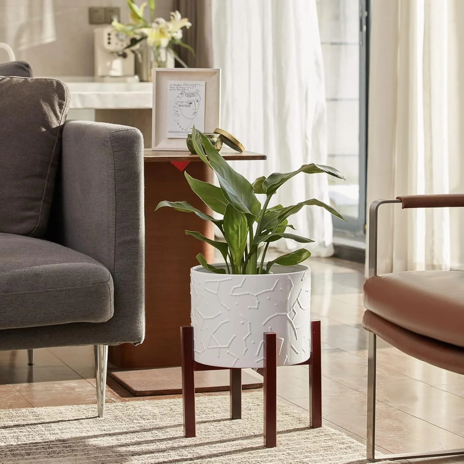 Indoor living room with a potted plant on a wooden stand, grey sofa, brown chair, side table with framed artwork, flowers, and a window with curtains in the background.