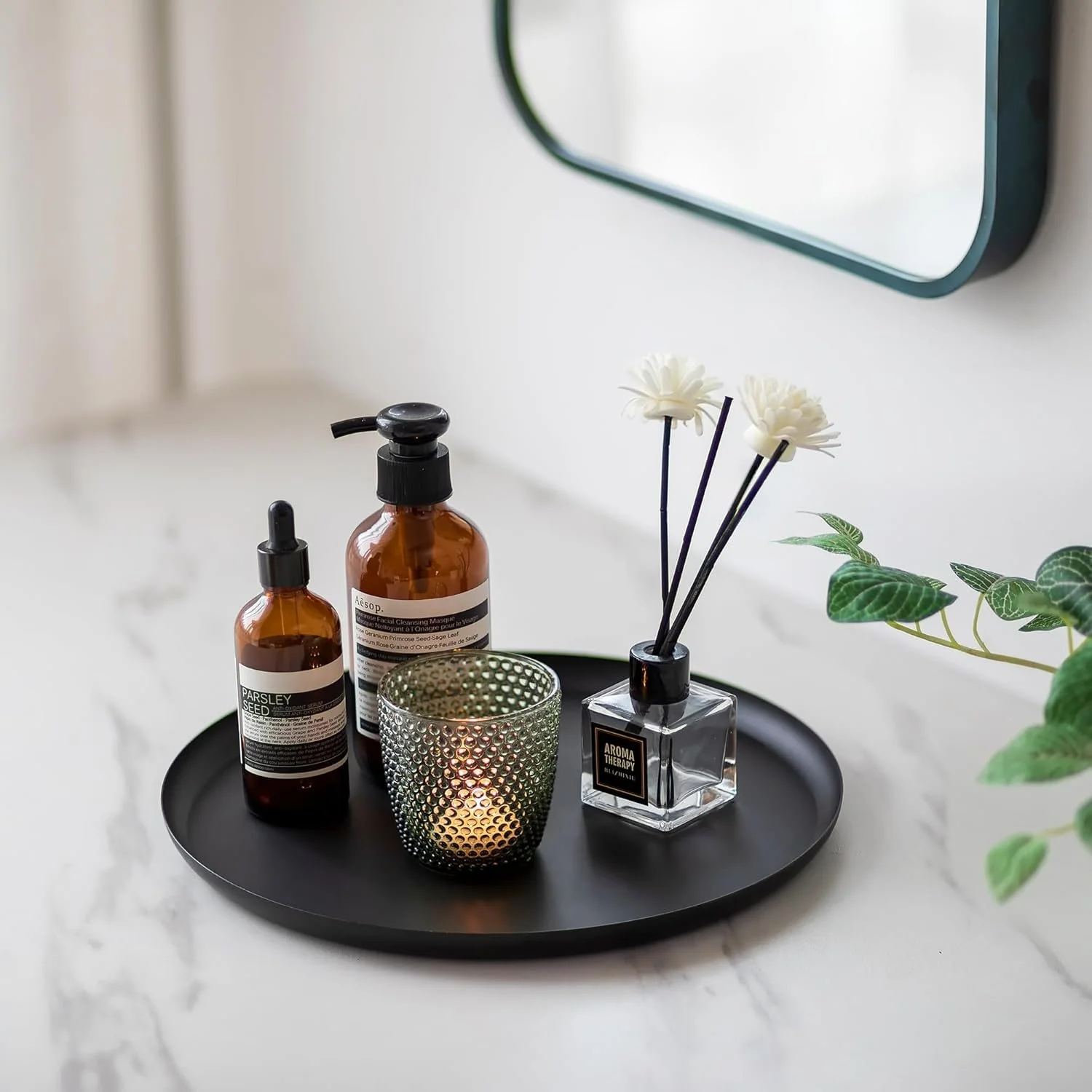 A black tray holding amber bottles labeled 'Parsley Seed' and 'Aesop', a glass candle holder with a lit candle, a vase with white artificial flowers, and a small potted green plant, placed on a white marble surface near a mirror.