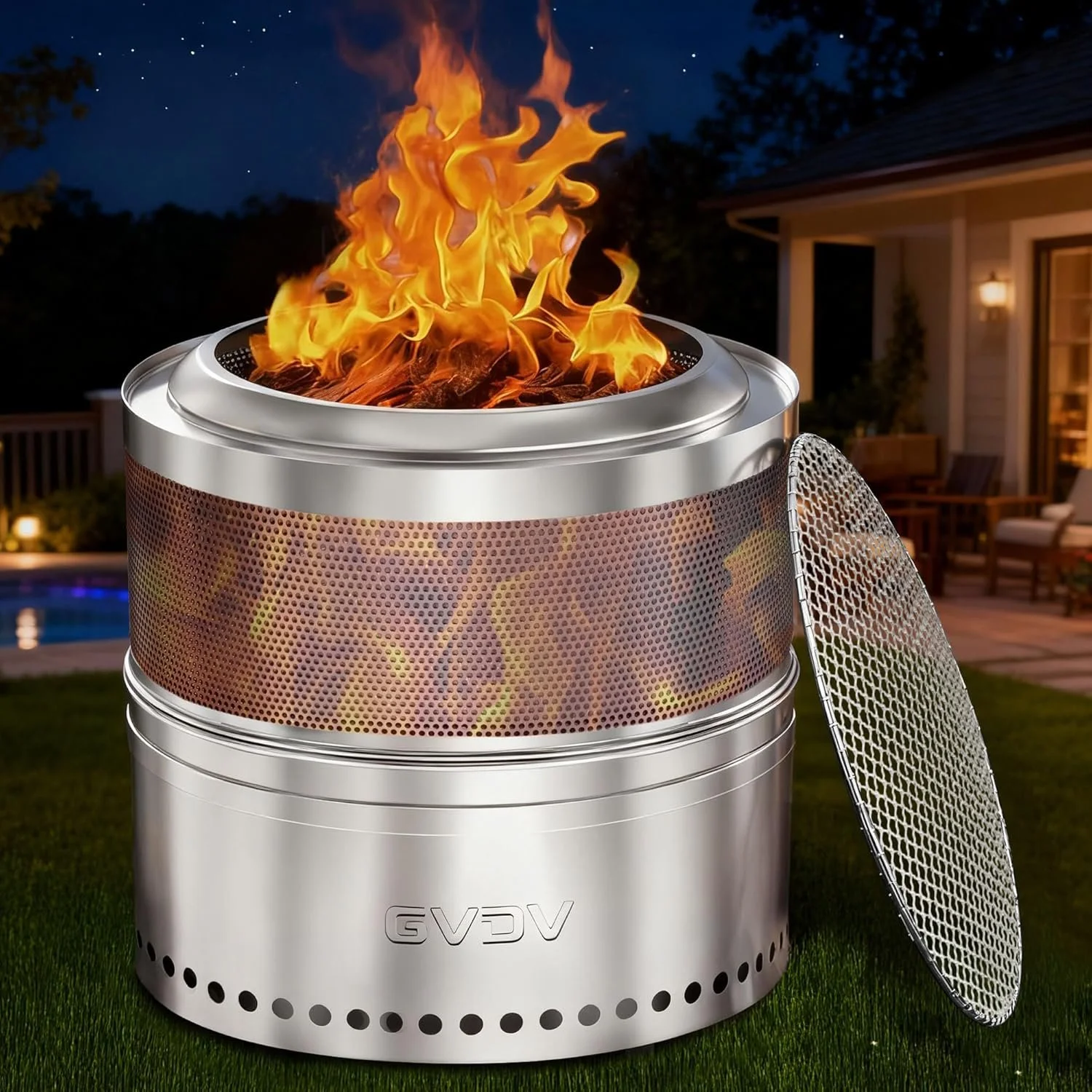 A modern stainless steel outdoor fire pit with orange and yellow flames, set in a backyard at night with a house and patio furniture in the background.