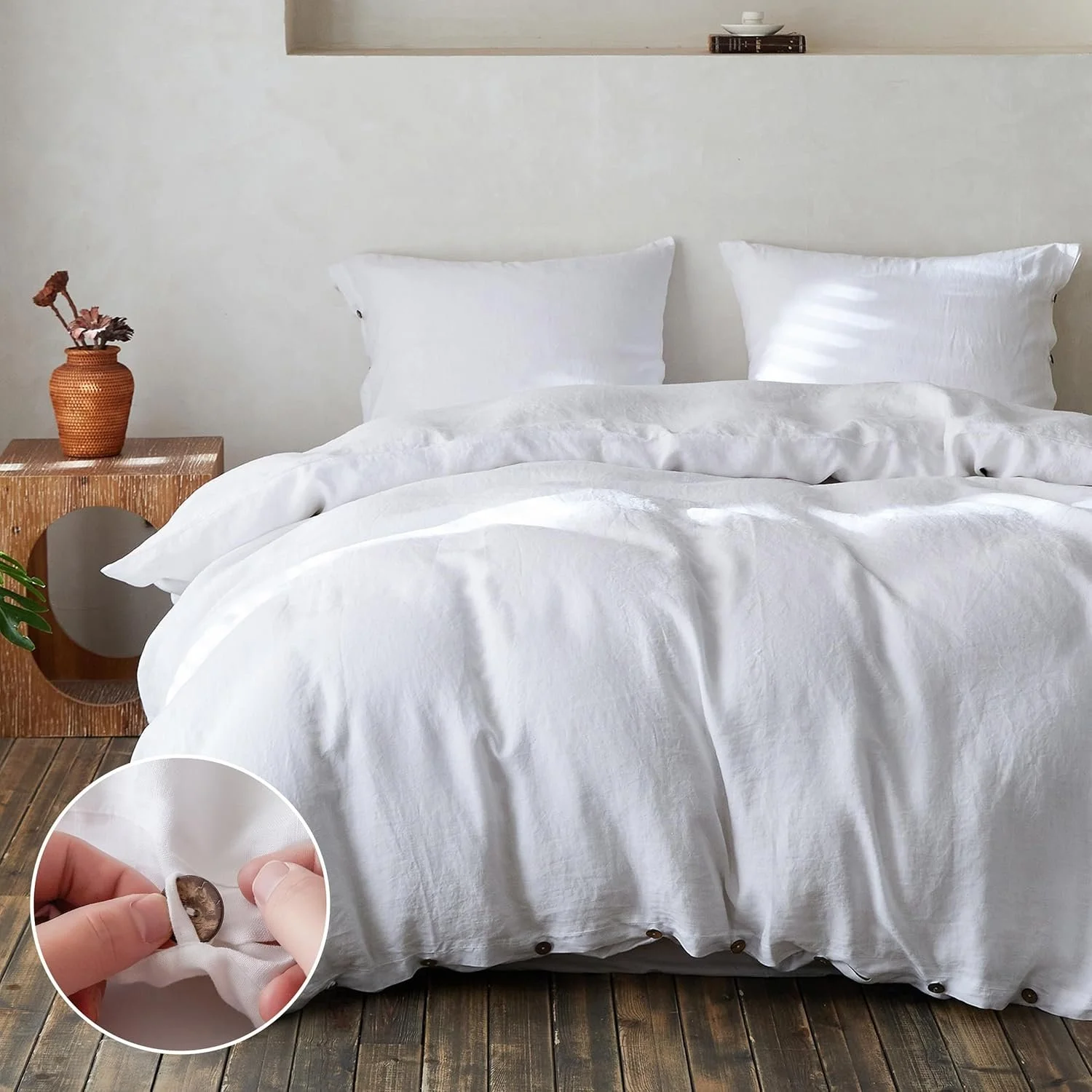 Close-up of a hand inserting a small coin into a hidden pocket inside a white duvet on a bed, with white pillows and a wooden bedside table with a woven vase and dried flowers, on a wooden floor in a minimalist bedroom.