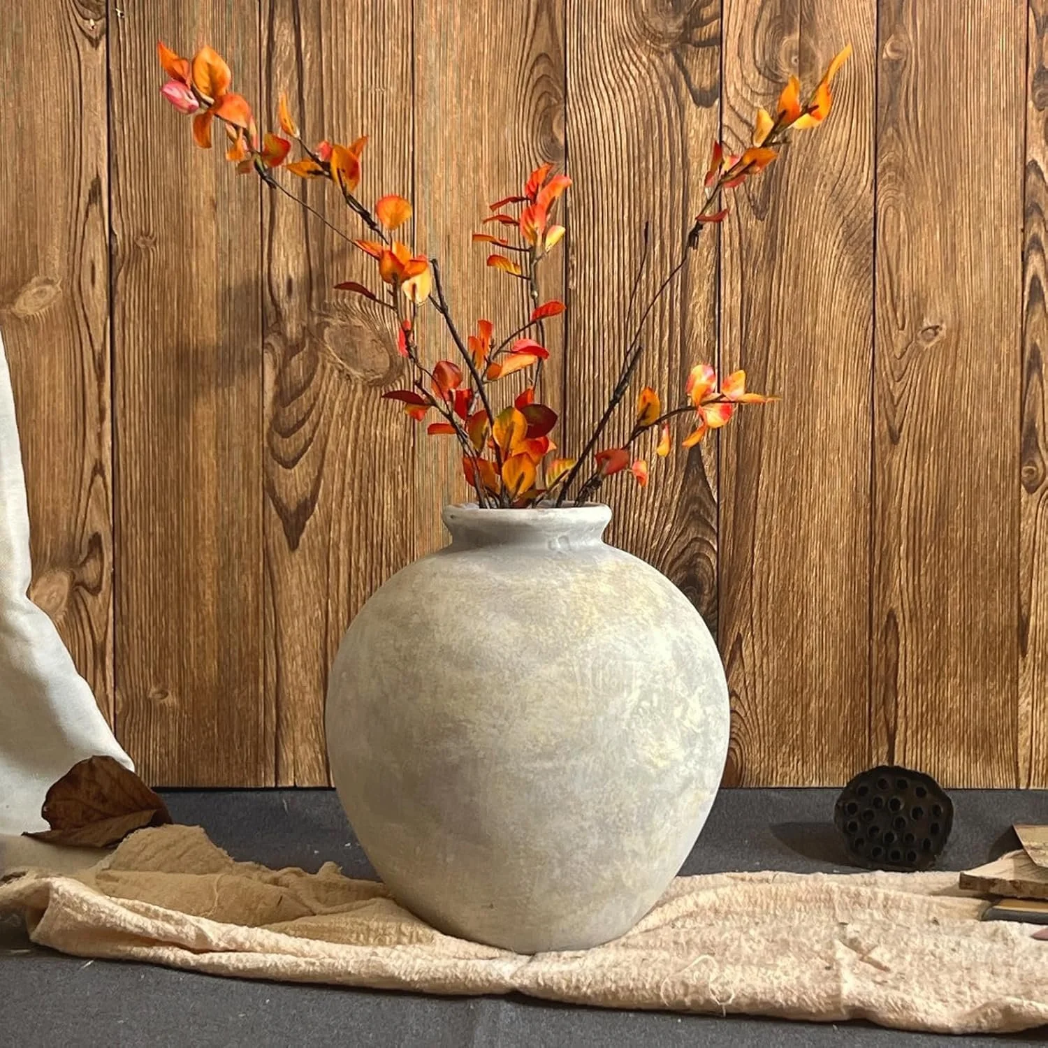 A round, beige ceramic vase with narrow opening holding orange and yellow artificial flowers, placed on a beige cloth on a gray surface against a wooden panel background.