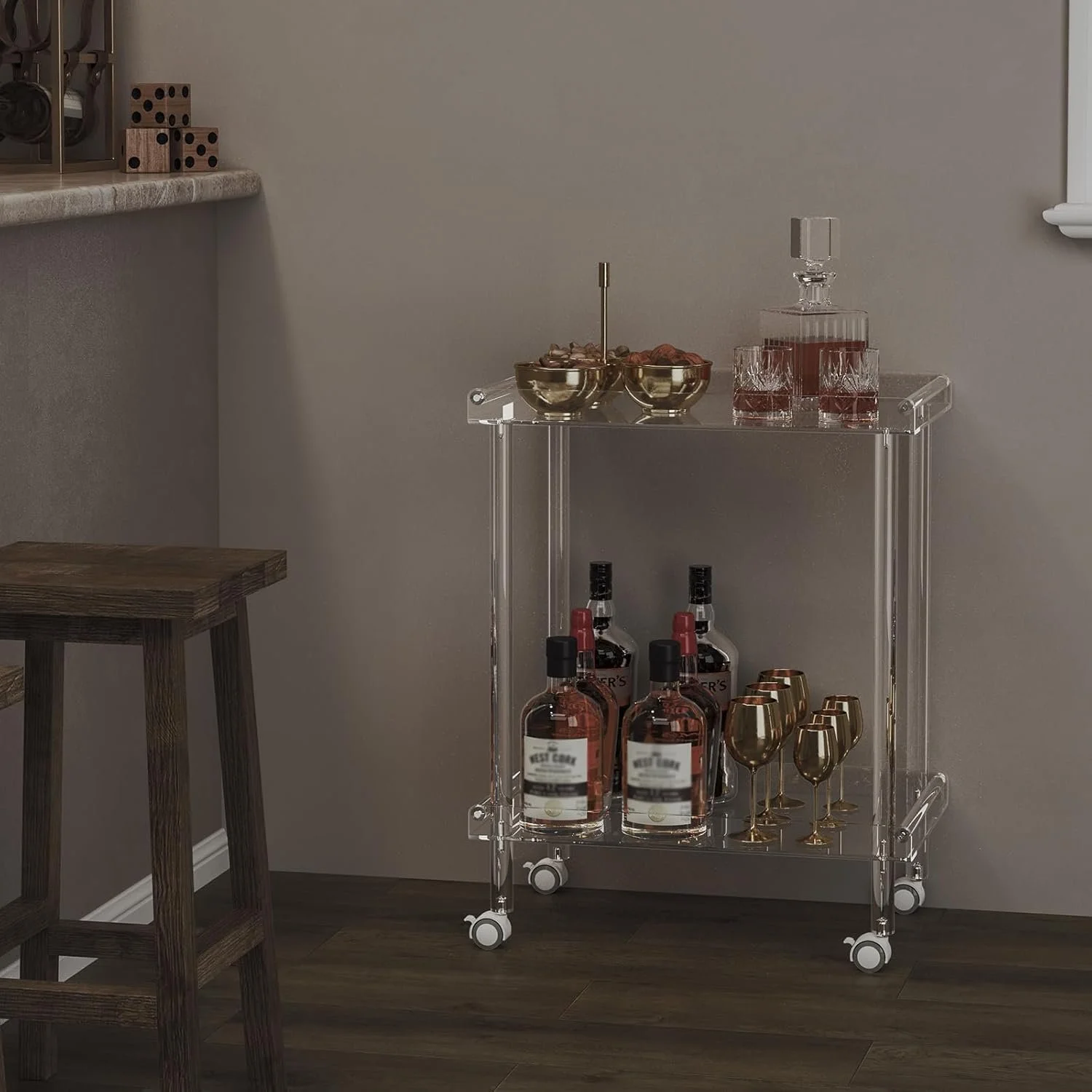 A clear acrylic bar cart on wheels with two shelves, holding bottles of alcohol, gold and glassware, and bowls of snacks in a room with brown hardwood floors and a neutral-colored wall.