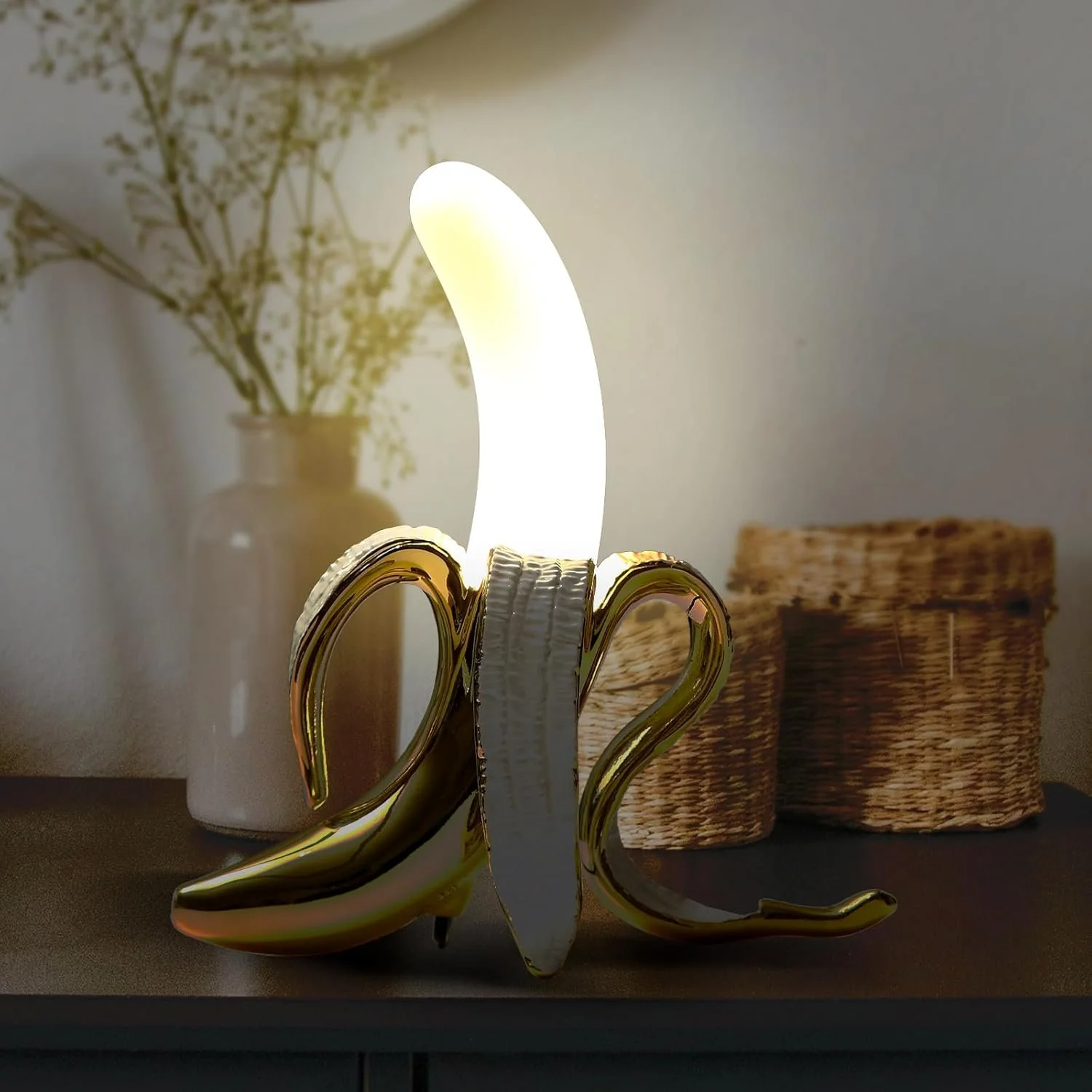 Decorative banana-shaped lamp with white and gold accents, lit and placed on a dark surface, with woven baskets and a glass vase with dried flowers in the background.