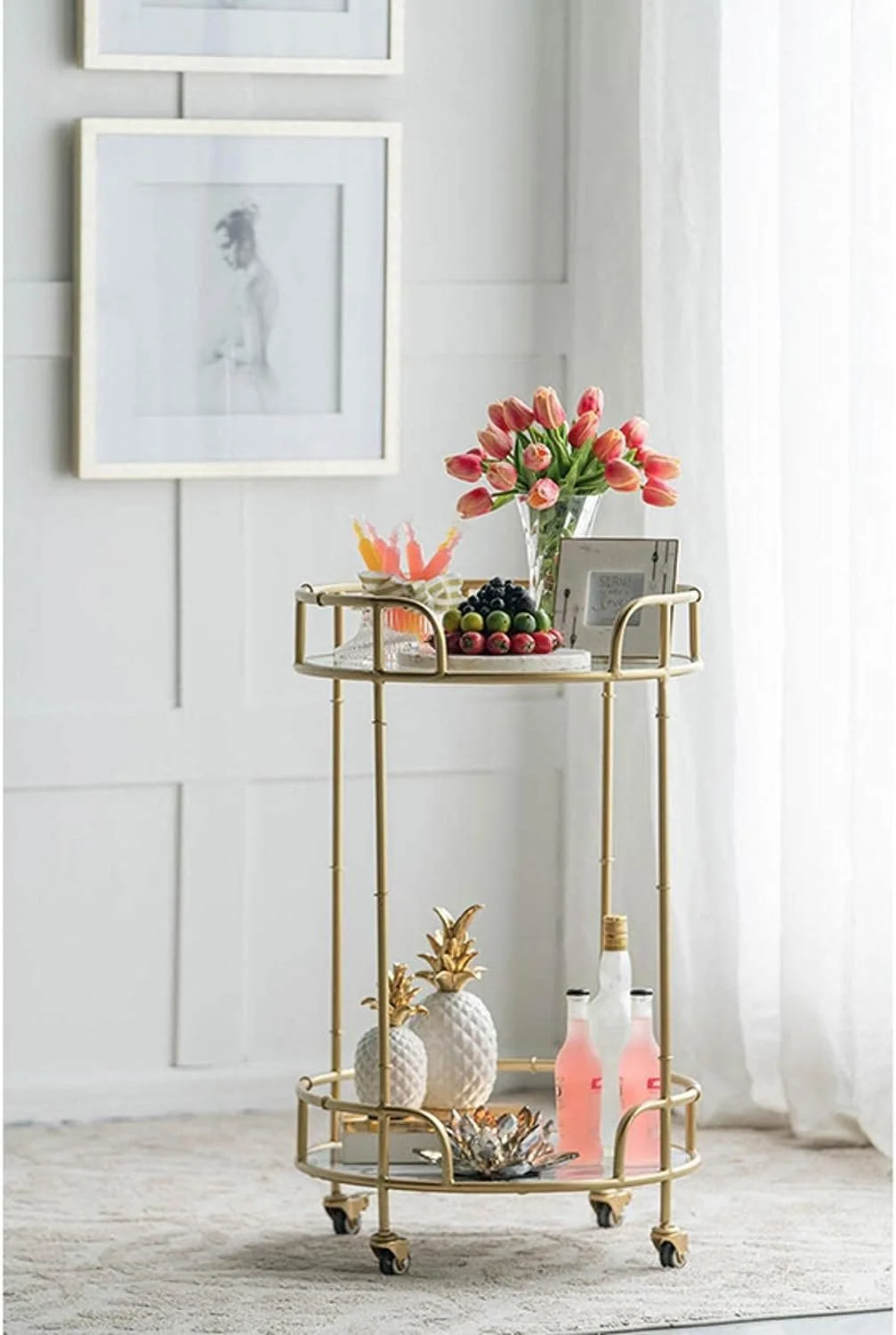 A gold bar cart with wheels holding a vase of pink and peach tulips, a platter of mixed berries, small decorative pineapples, and pink bottles. Behind the cart is a white wall with two framed photographs, and a sheer white curtain to the right.