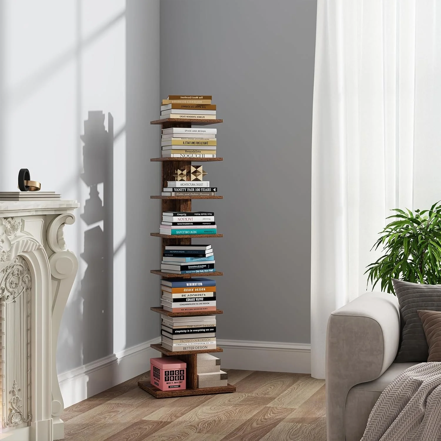 A tall wooden bookshelf filled with books is placed against a gray wall in a living room. To the right, a window with sheer white curtains, a green potted plant, and part of a beige sofa with pillows are visible.
