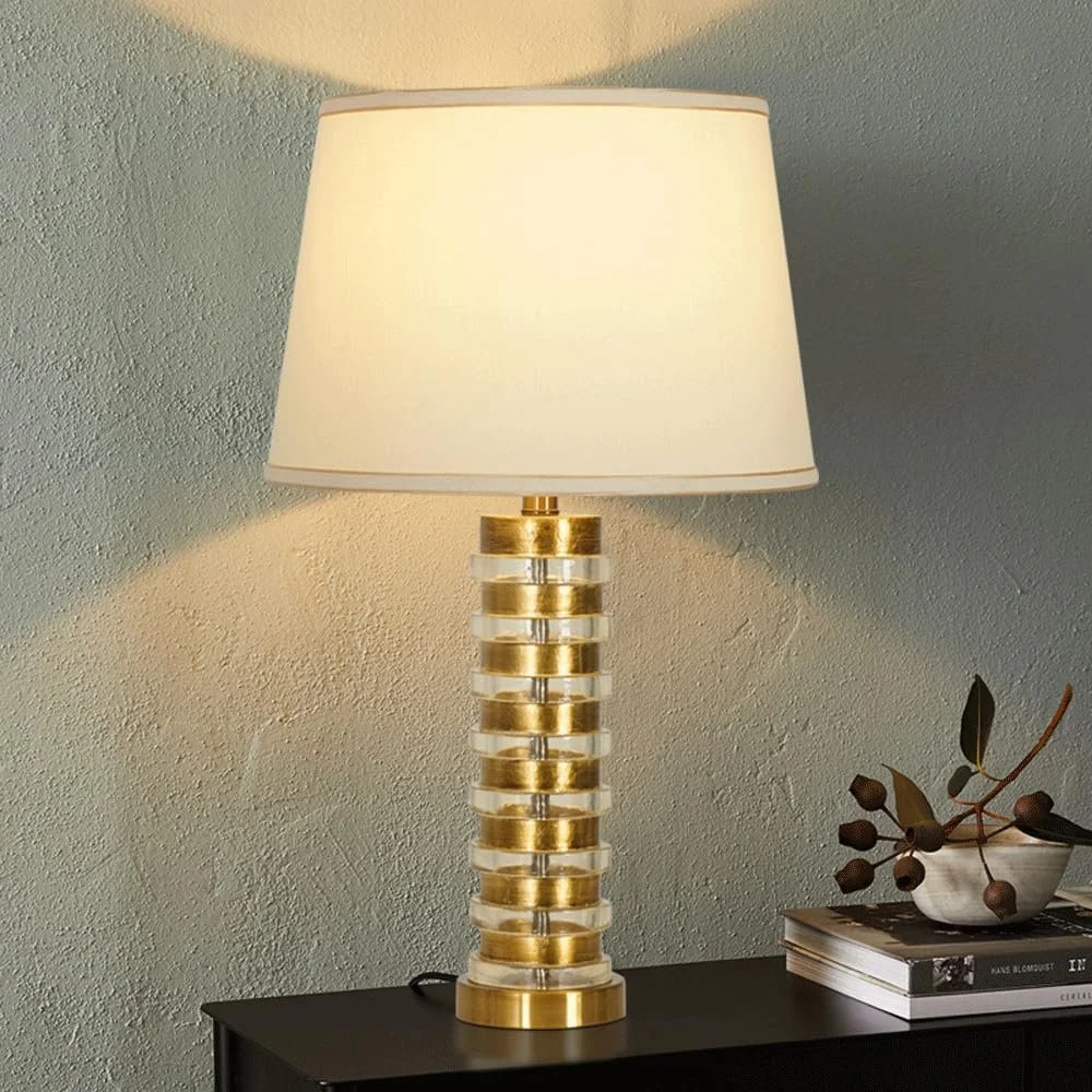 A decorative table lamp with a cylindrical base featuring alternating gold and transparent rings, resting on a black table next to a stack of books and a bowl with branches and round dark brown decorations.