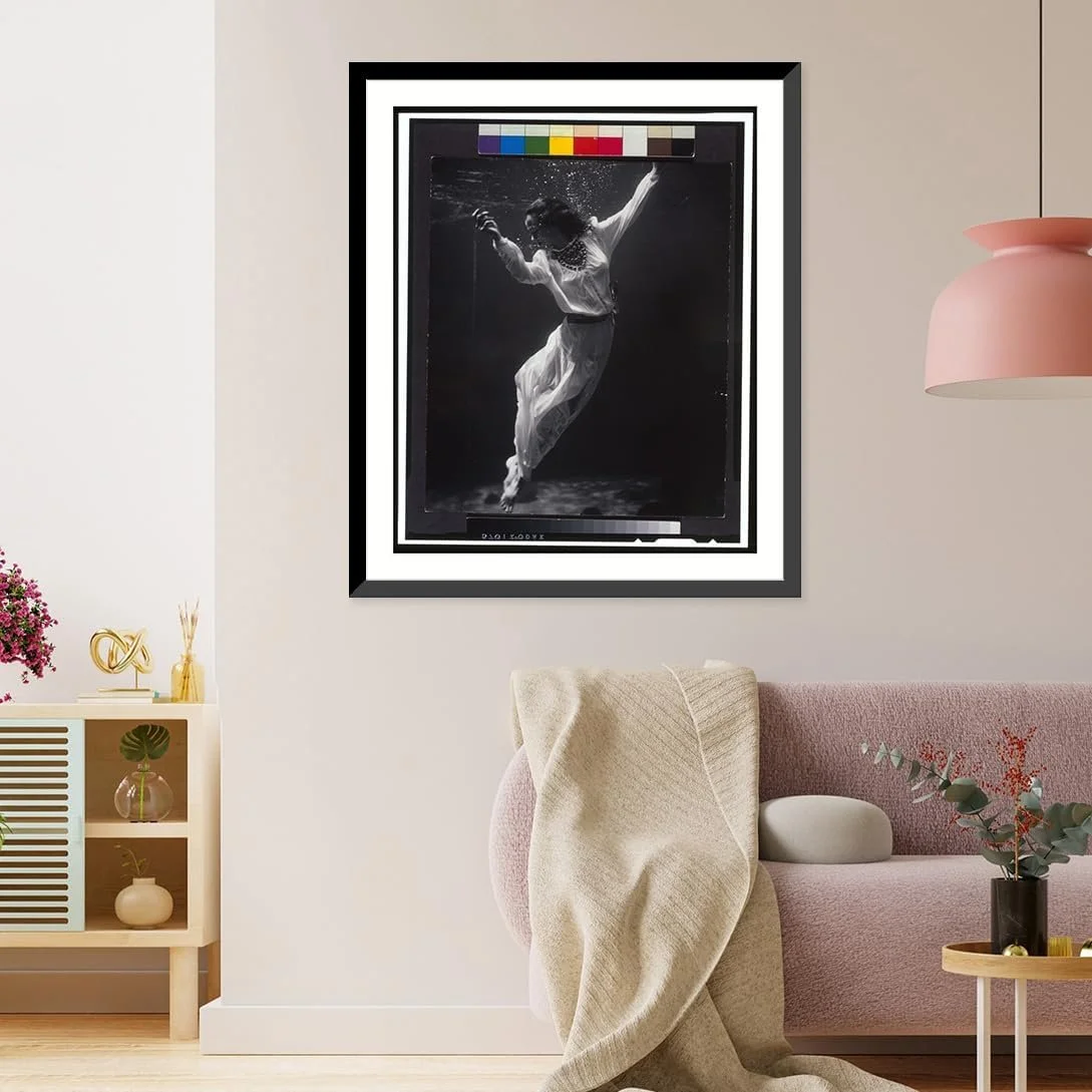 A black-and-white framed photograph of a woman in a flowing dress underwater, hanging on a wall in a modern living room with pink and neutral decor.