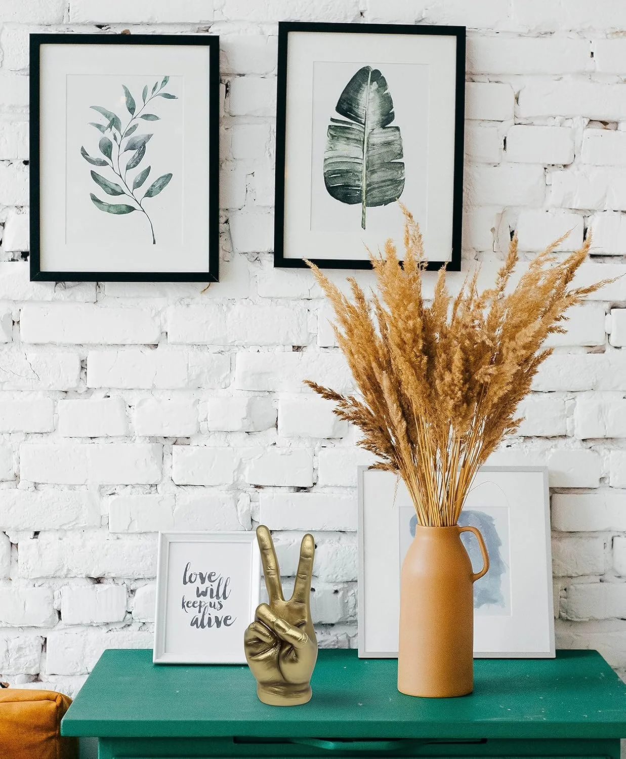 Decorative display with framed botanical prints, a gold hand-shaped sculpture making a peace sign, a framed quote reading 'love will keep us alive', a large beige vase with dried pampas grass, and a green table against a white brick wall.