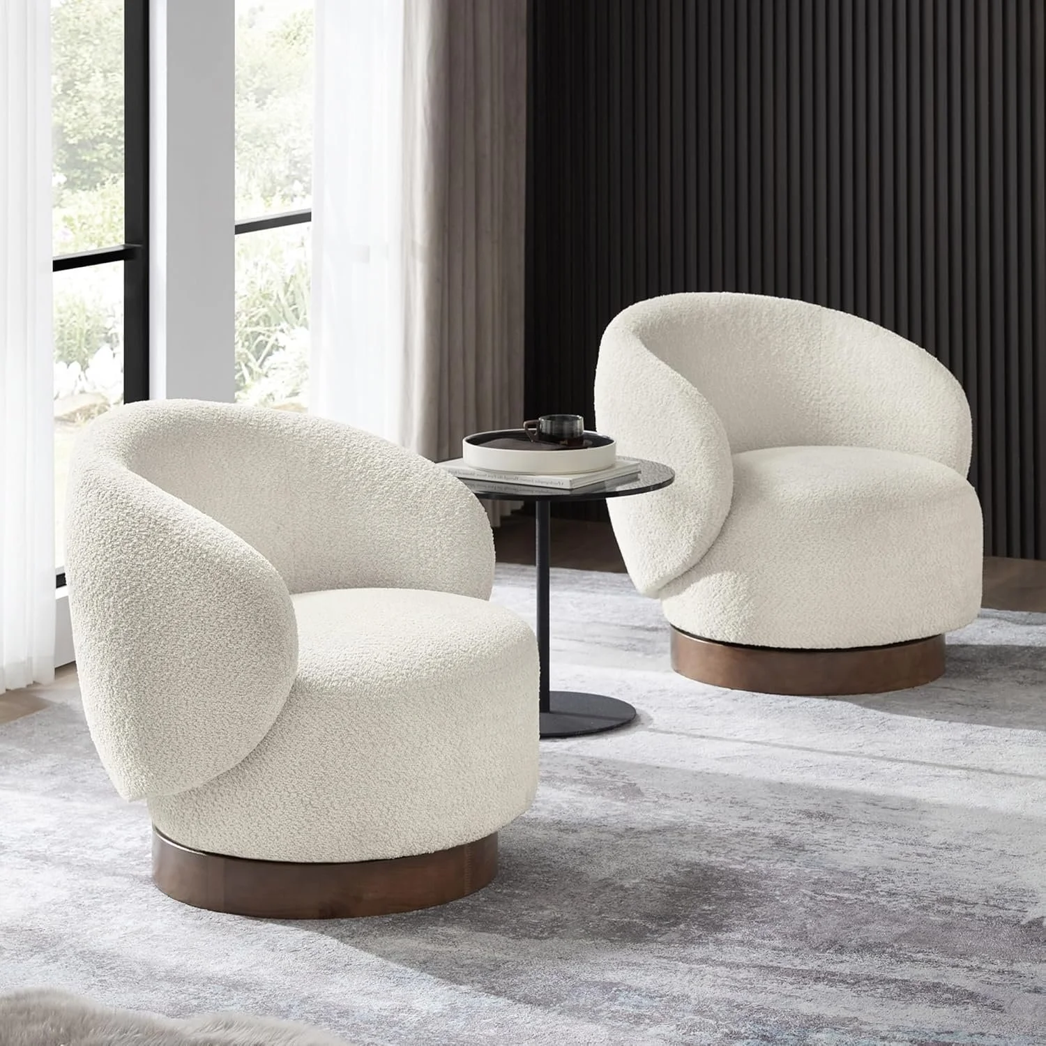 Two modern cream-colored boucle armchairs with rounded backs and dark wooden bases, separated by a black round side table with a tray and cup, in a bright room with large windows, sheer curtains, and a dark textured accent wall.