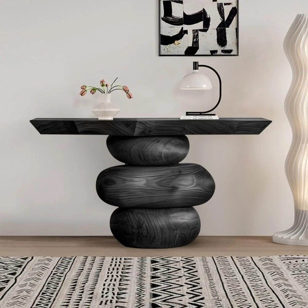A black sculptural wooden console table with rounded base supports, topped with a white table lamp, a stack of books, and a small vase with flowers, against a white wall with black and white abstract wall art and a beige patterned rug on the floor.