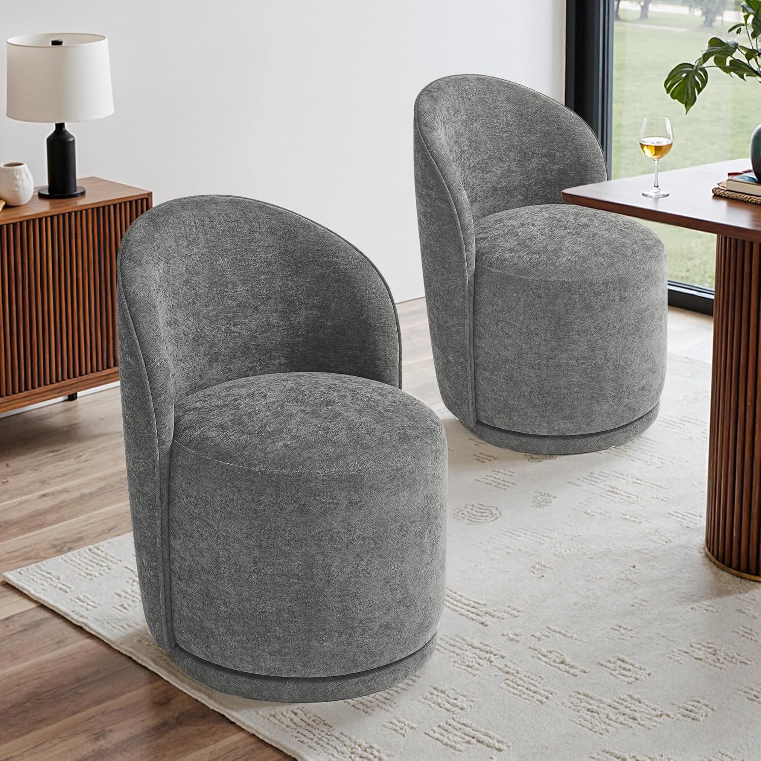 Two modern gray upholstered armchairs in a bright room with wooden floors, a white rug, a wooden sideboard with a black lamp, and a window with greenery outside.