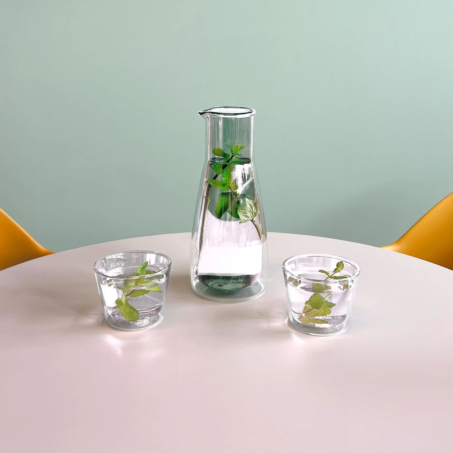 Glass water pitcher with leafy mint and basil immersed, on a white table flanked by two glasses containing leaf-infused water and mint and basil leaves inside.