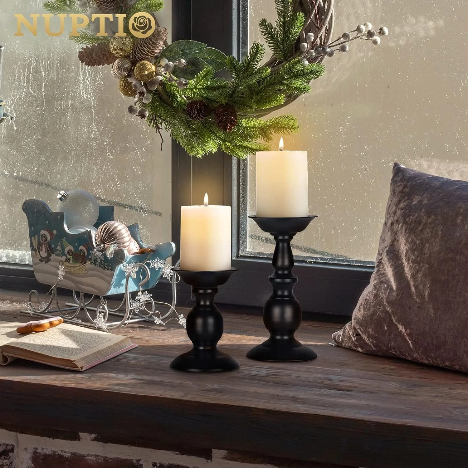 A cozy holiday scene featuring two lit white candles on black candlesticks, a velvet pillow, an open book with a pen on top, a decorative sleigh with Christmas ornaments, and a mistletoe wreath hanging in front of a rain-streaked window.