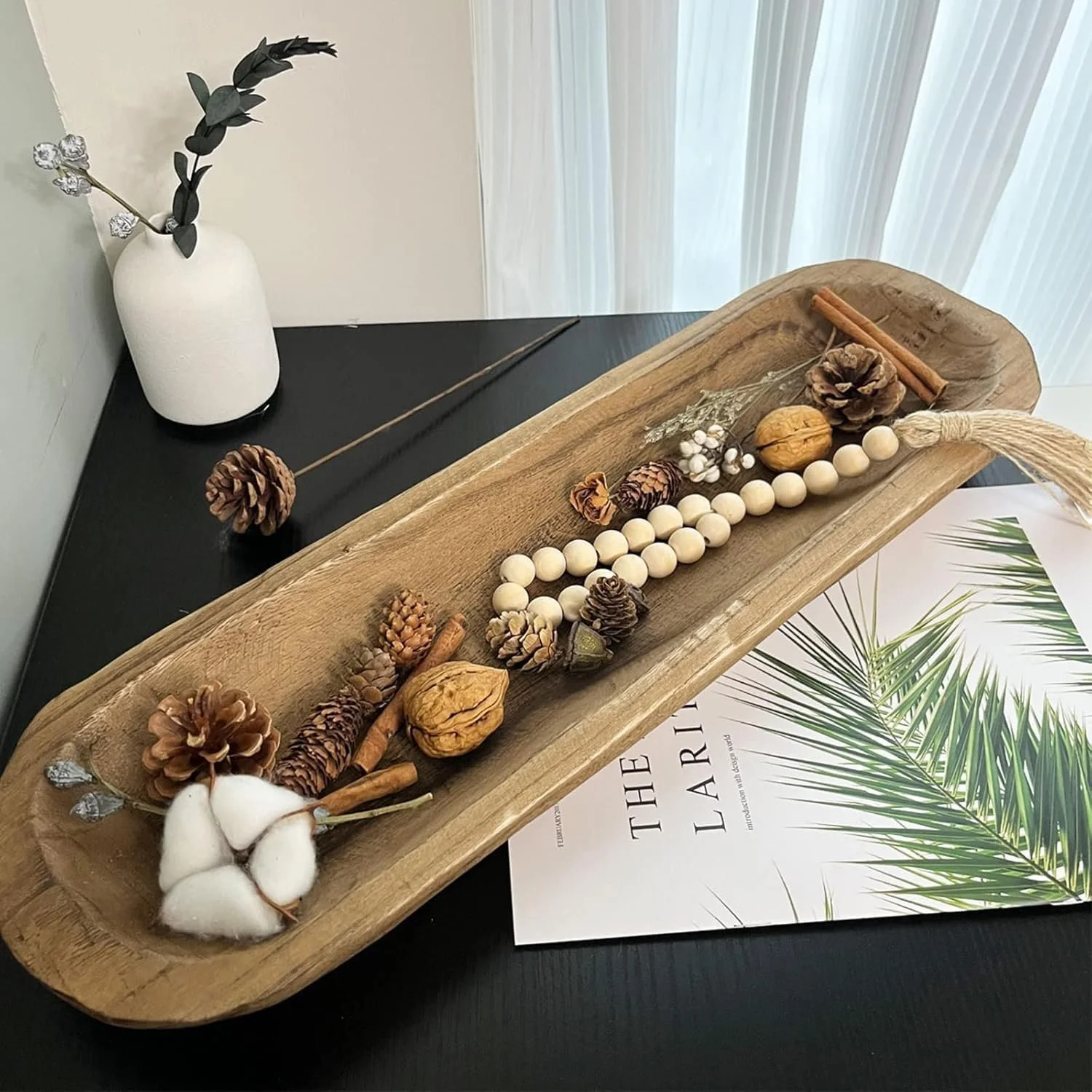 Decorative wooden tray with pinecones, cinnamon sticks, walnuts, white beaded string, and cotton on a black surface, with a white vase and artificial plant in the background.
