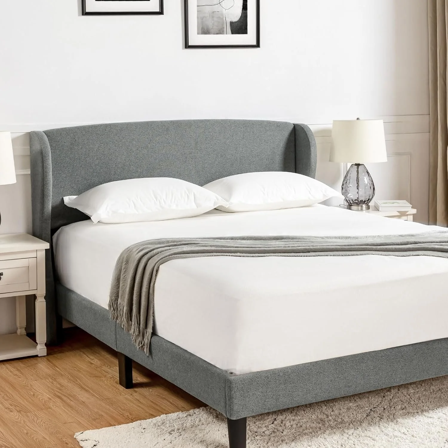 A neatly made bed with a gray upholstered headboard, white sheets, two white pillows, and a beige throw blanket, with nightstands and table lamps on either side in a minimalist bedroom.