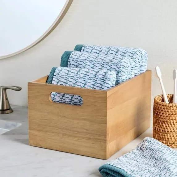 A wooden box filled with folded blue and white patterned towels on a bathroom countertop, with a wicker basket holding toothbrushes and a faucet in the background.