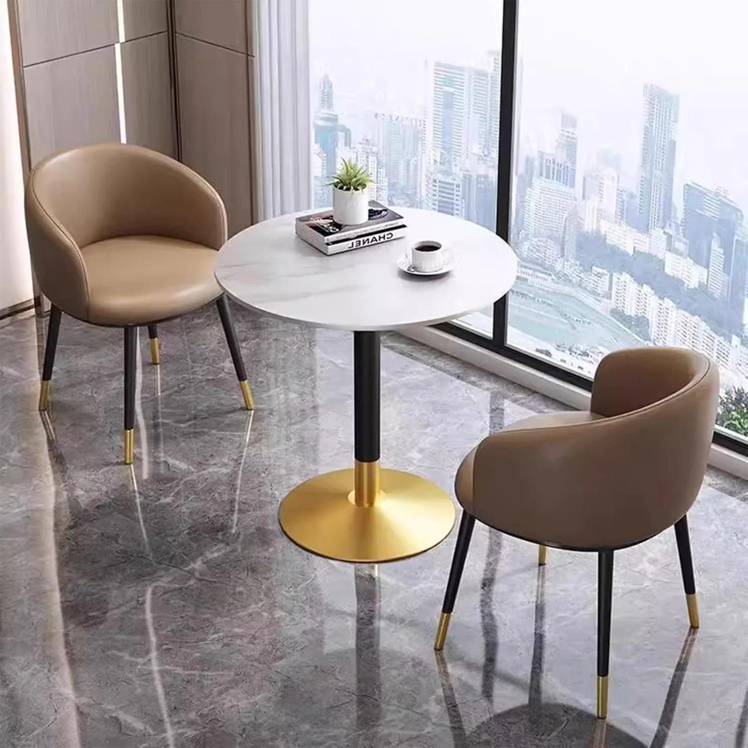 Round white marble table with two beige armchairs with black legs and gold tips, located in front of large windows showing a cityscape view.