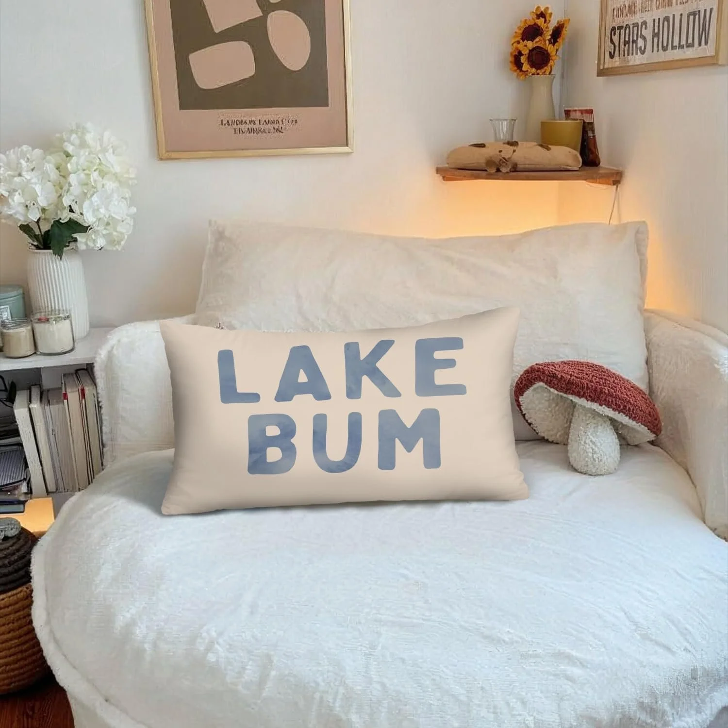 Decorated bedroom with a white bed, white pillows, a pillow with the text 'LAKE BUM,' a mushroom-shaped plush toy, a shelf with sunflowers, framed artwork, and a side table with candles and books.