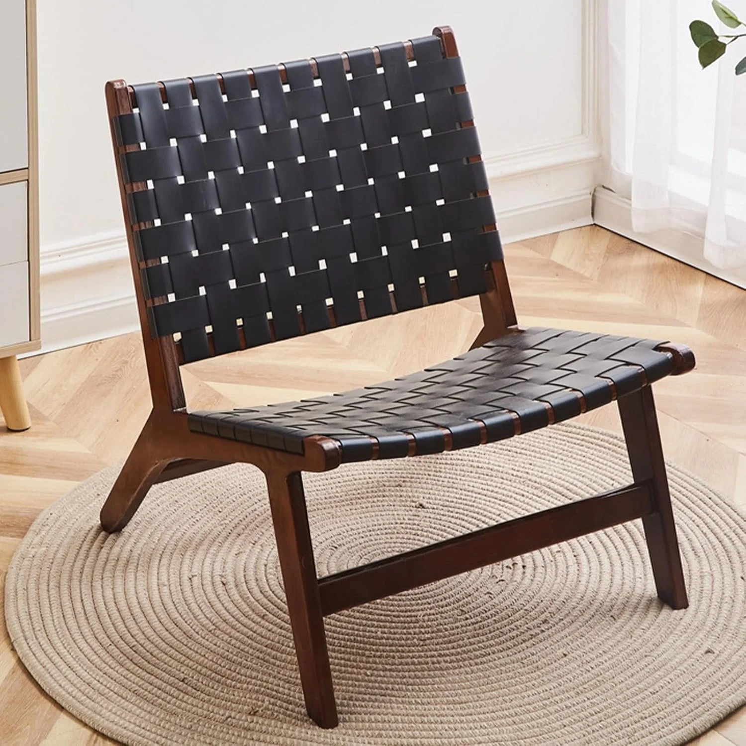 A modern wooden lounge chair with black woven straps, placed on a circular beige rug inside a bright room near a window with sheer white curtains.