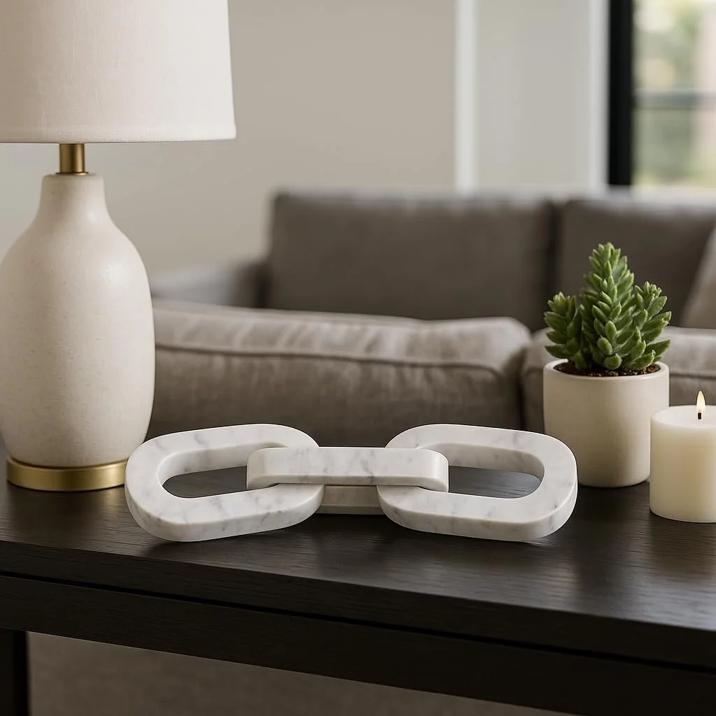 Decorative table with a white ceramic table lamp, a white marble chain link sculpture, a potted succulent, and a white candle, in a modern living room.