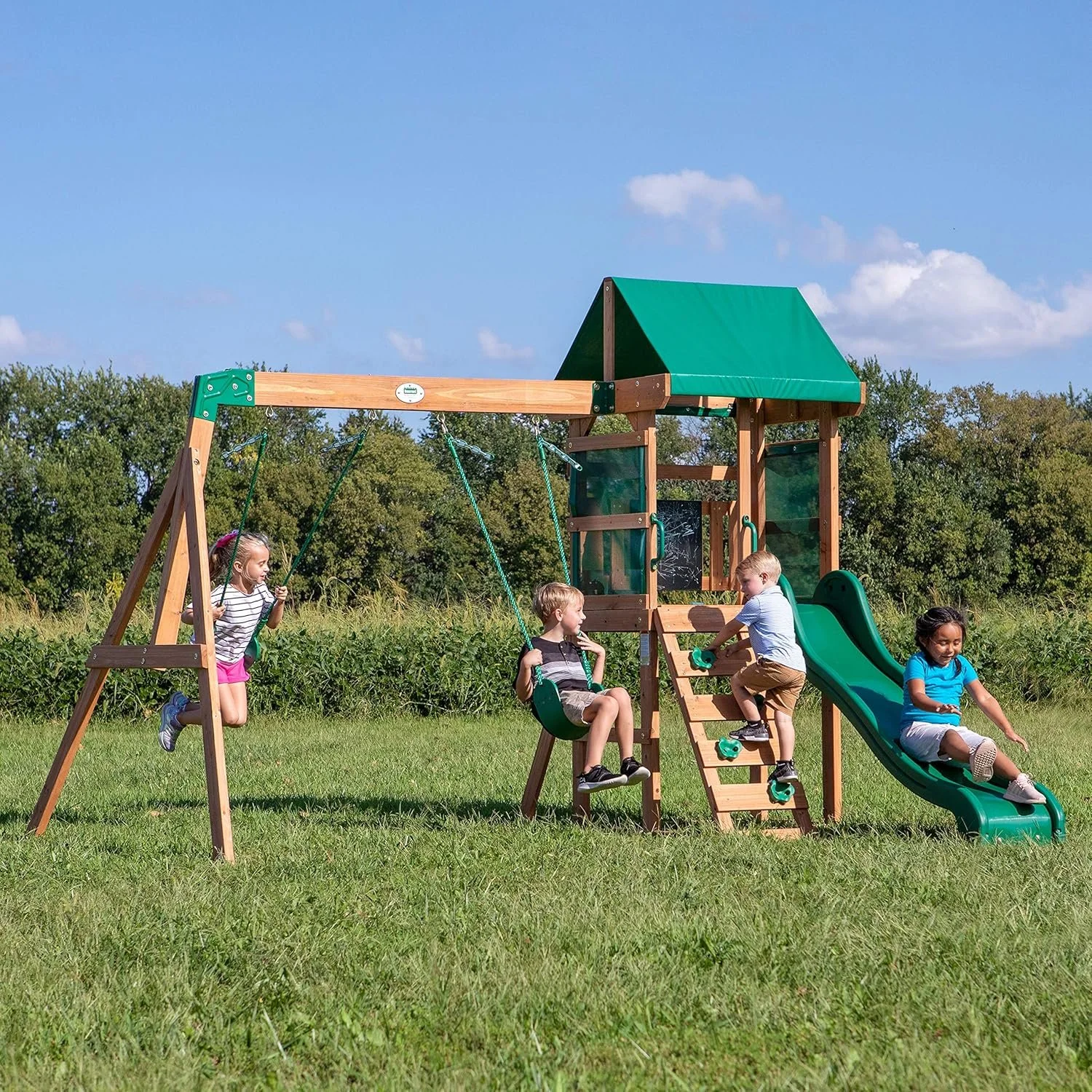 Children playing on a wooden playground set with swings, slides, and climbing features in an open grassy field under a blue sky.