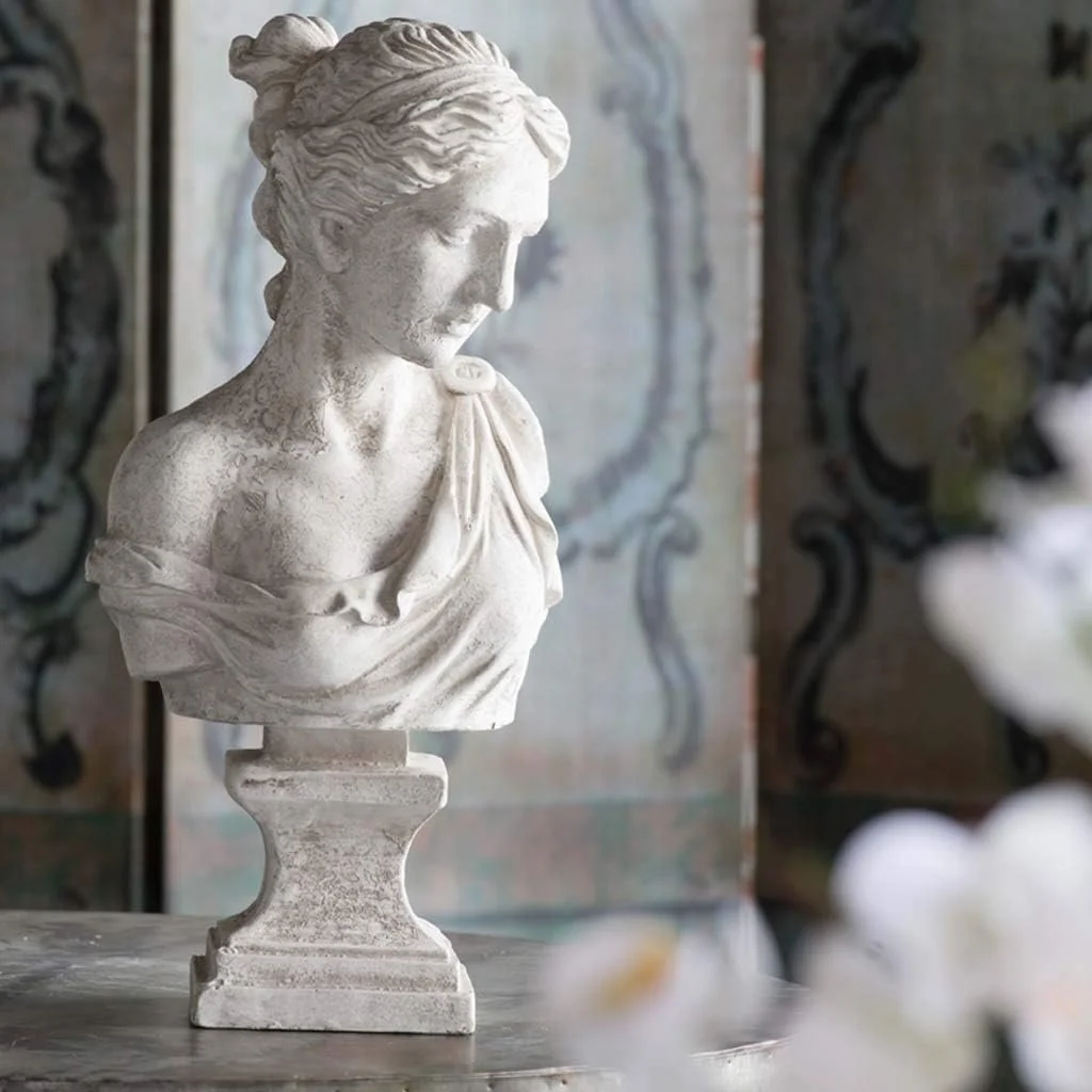 A white marble bust sculpture of a woman with her hair tied up, looking downward, placed on a pedestal. The background features decorative curtains or panels with swirling patterns.