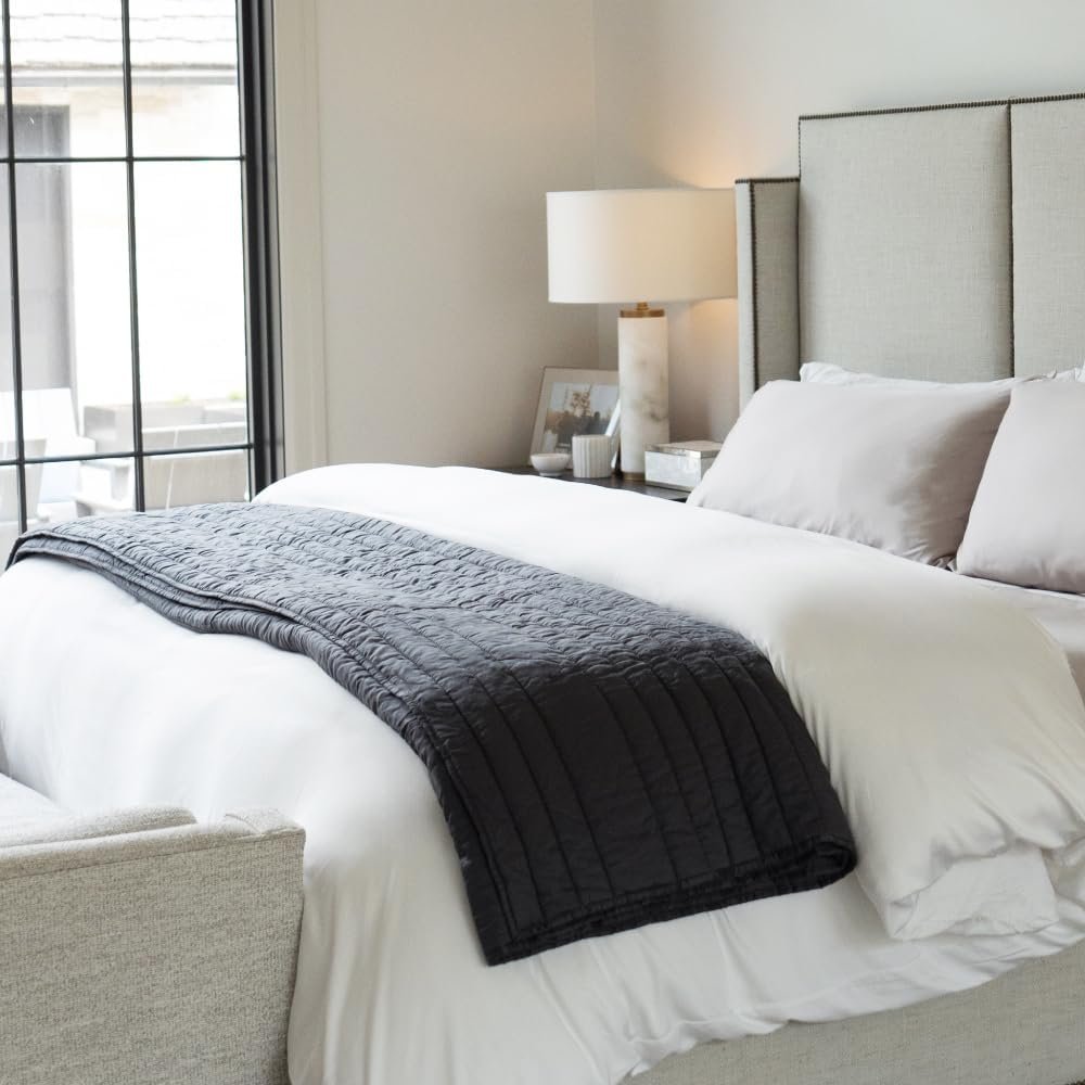 A neatly made bed with white sheets, a black quilted blanket at the foot, and beige pillows, in a contemporary bedroom with a bedside table, lamp, and framed photo, next to a large window.