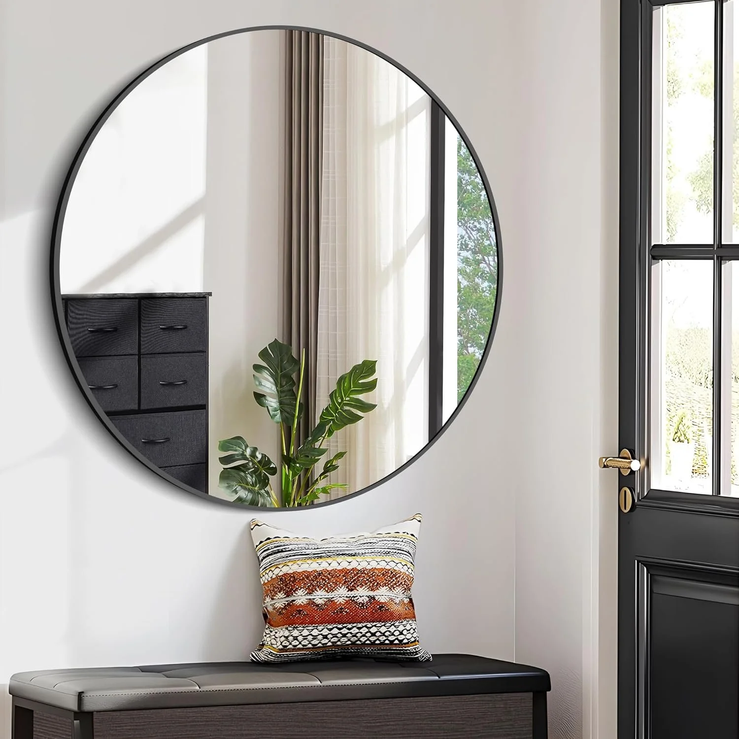 A modern entryway with a large round mirror, a black bench with a patterned pillow, a black dresser, a houseplant, sheer curtains, and a window with black framing.