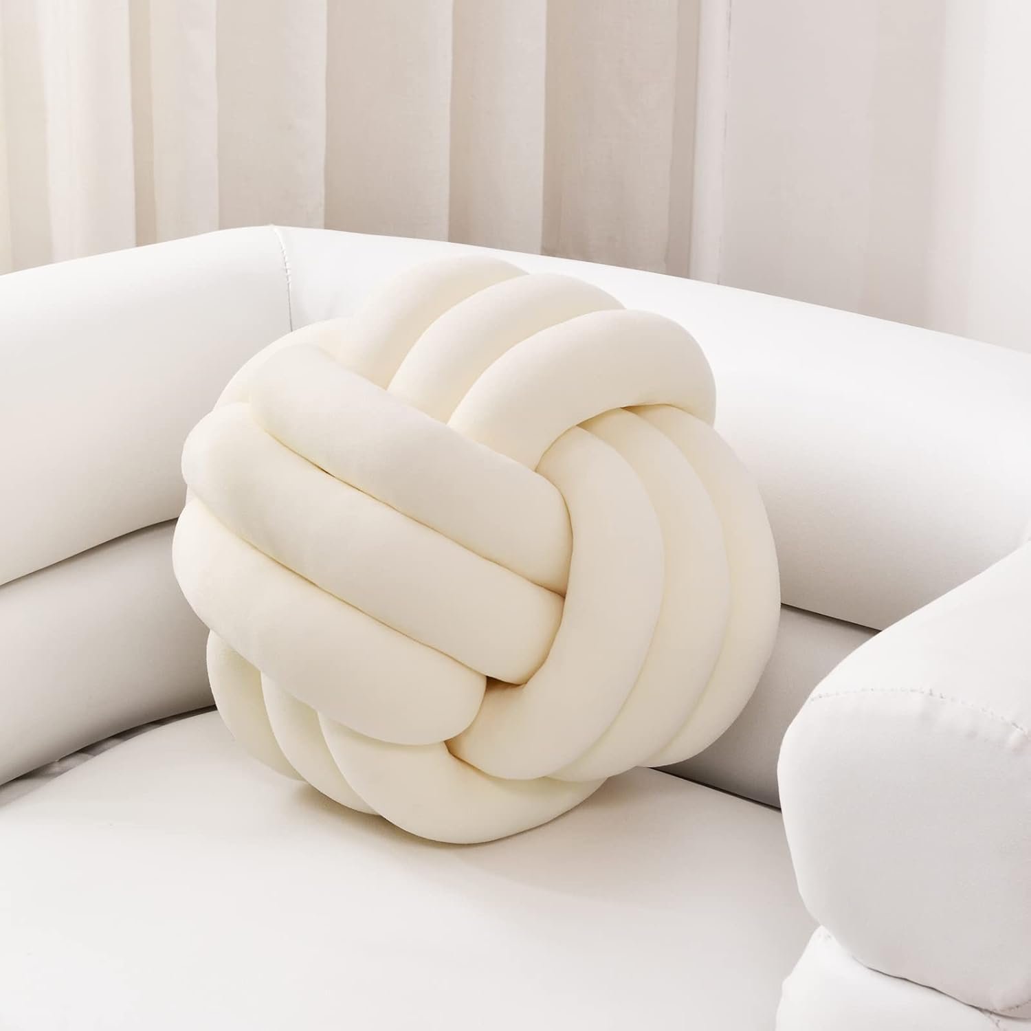 A white, knot-shaped cushion on a white modern sofa with light-colored vertical striped wall in the background.