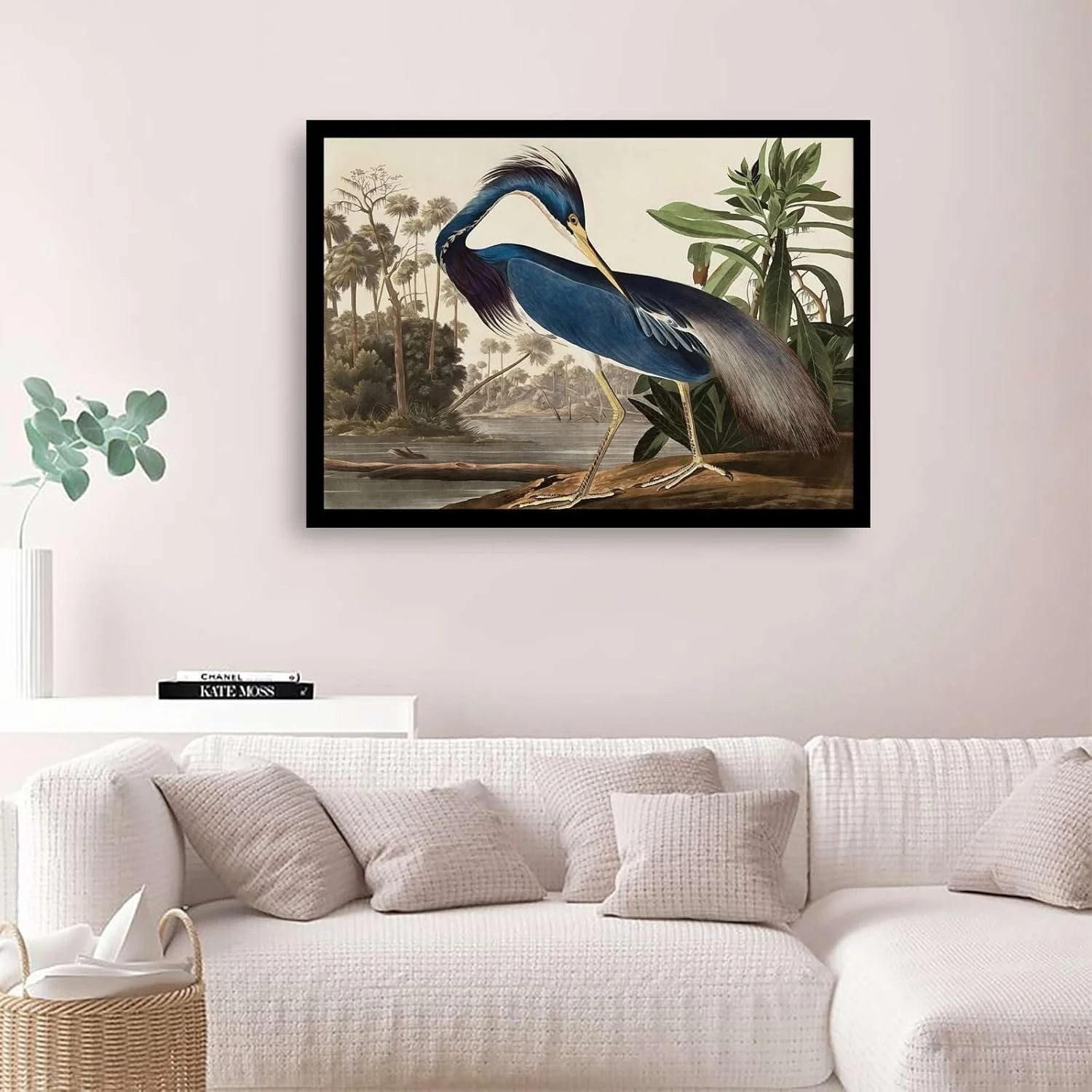 A framed picture of a heron standing by a river with trees and plants in the background, hanging on a light-colored wall above a white sofa with beige cushions.