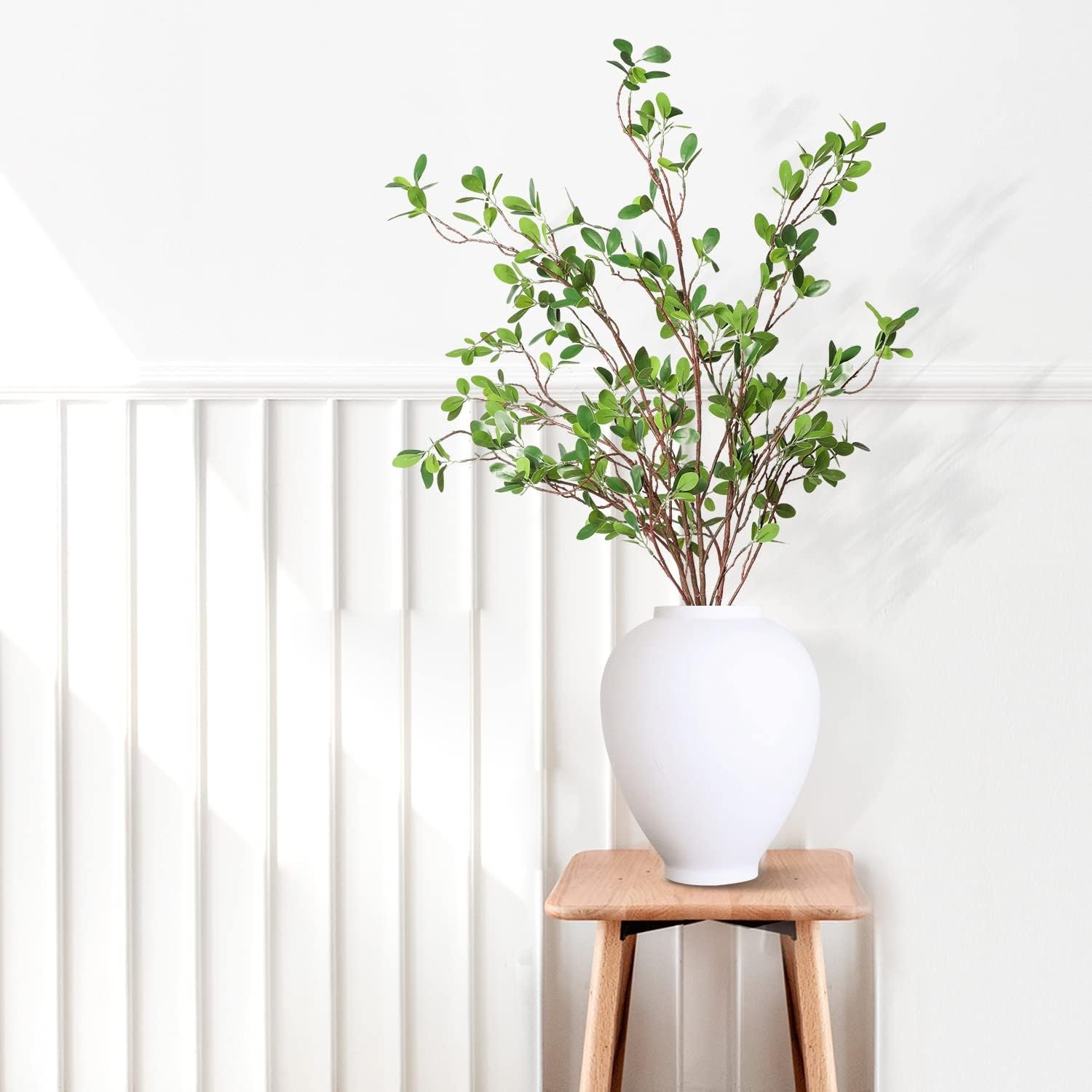 A white vase with green leafy branches on a wooden stool against a white wall.