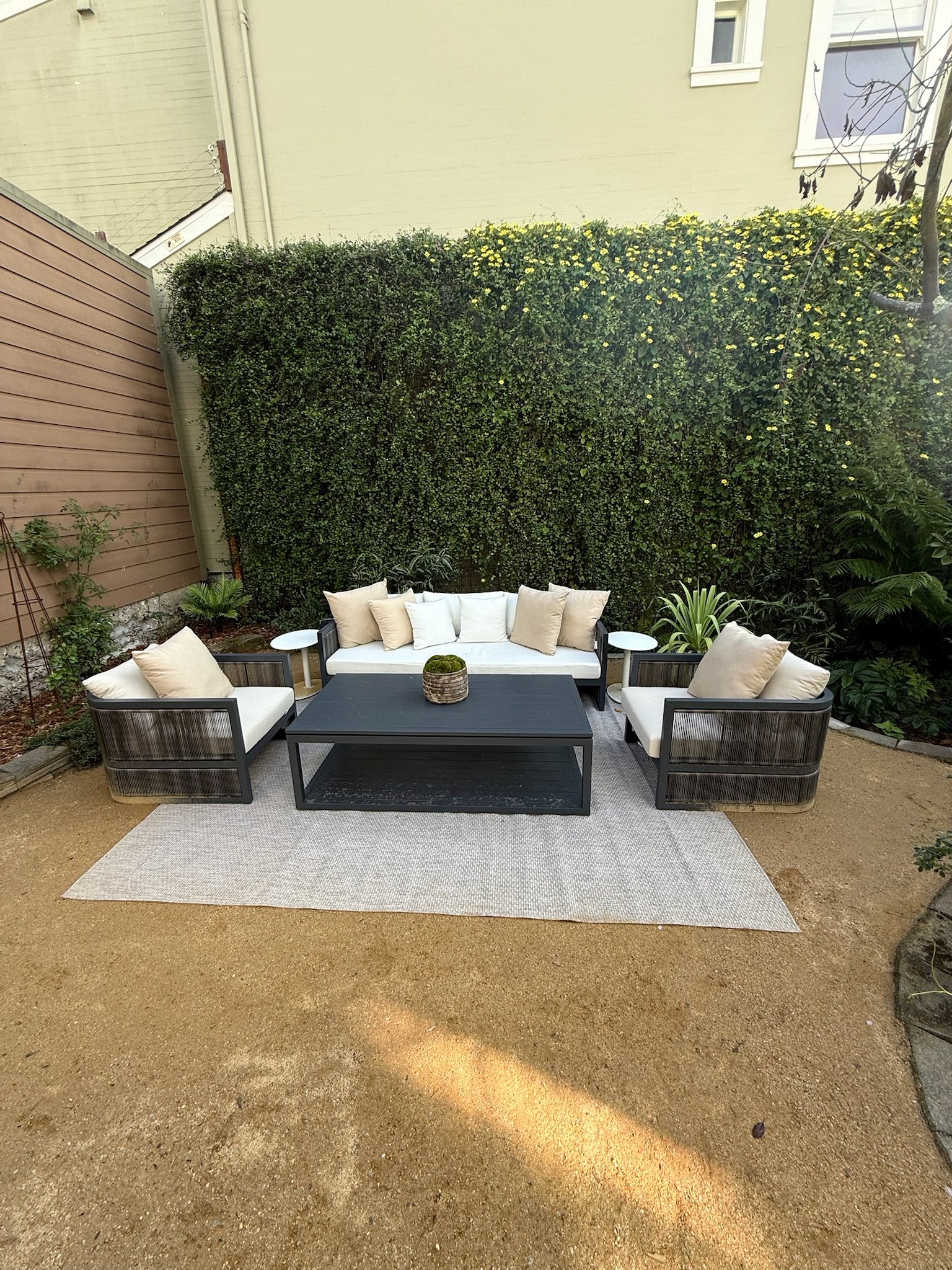 Outdoor patio area with seating furniture, including a sofa and two armchairs with light-colored cushions, a black coffee table, a white rug on a sandy ground, and a green hedge wall in the background.