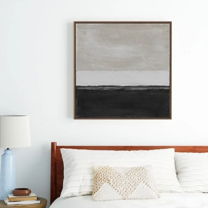 A bedroom with a framed abstract painting above the bed, a white bed with striped pillows, a wooden headboard, a blue and white table lamp, and a small brown dish and books on the nightstand.