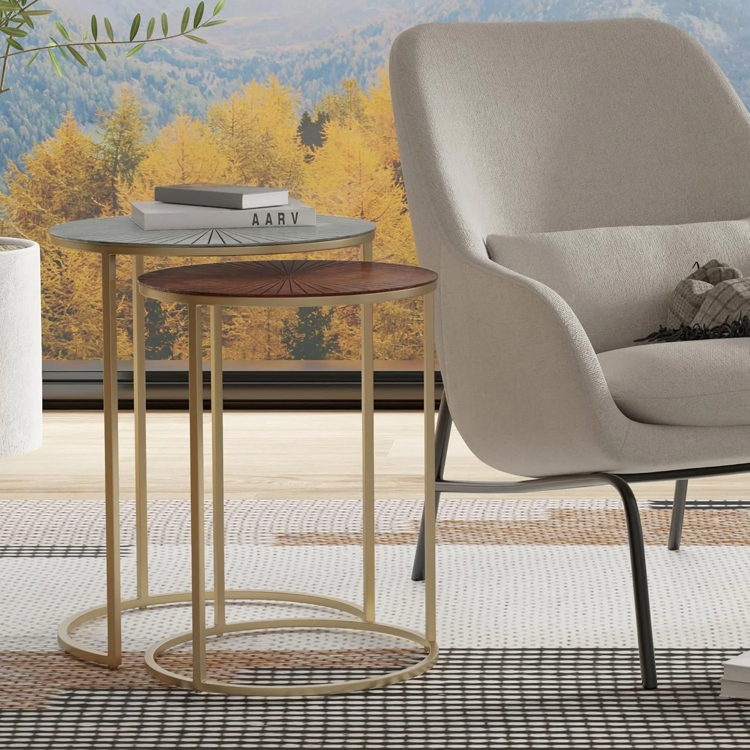 Two round nested side tables, one with a light wood top and the other with a dark wood top, placed beside a modern light-colored armchair with a gray metal leg. A wall mural of autumn trees with yellow and orange foliage in the background.