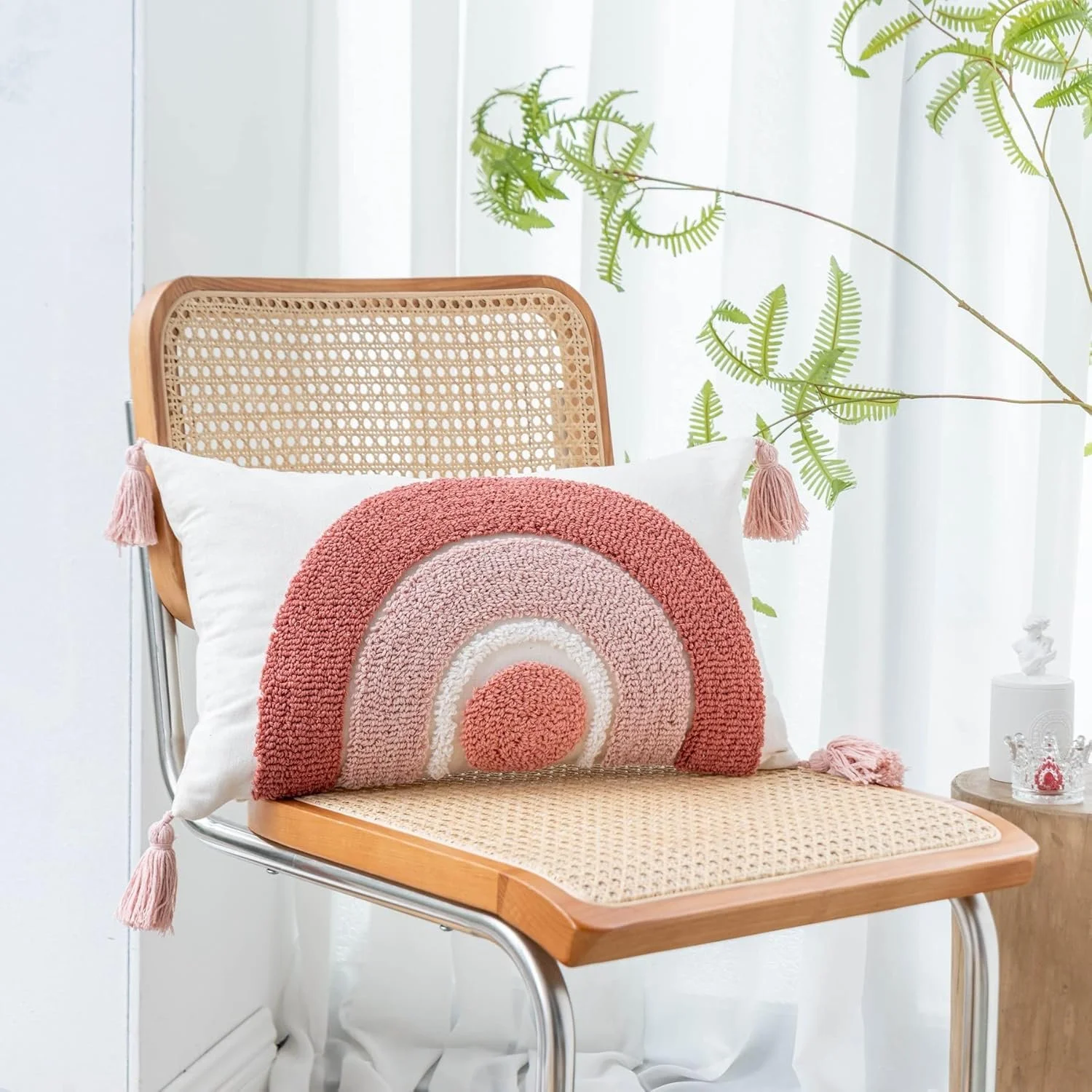 Rattan chair with a decorative rainbow pillow with tassels, placed in front of a white curtain and green fern plants, on a light-colored bamboo side table with small decorative items.