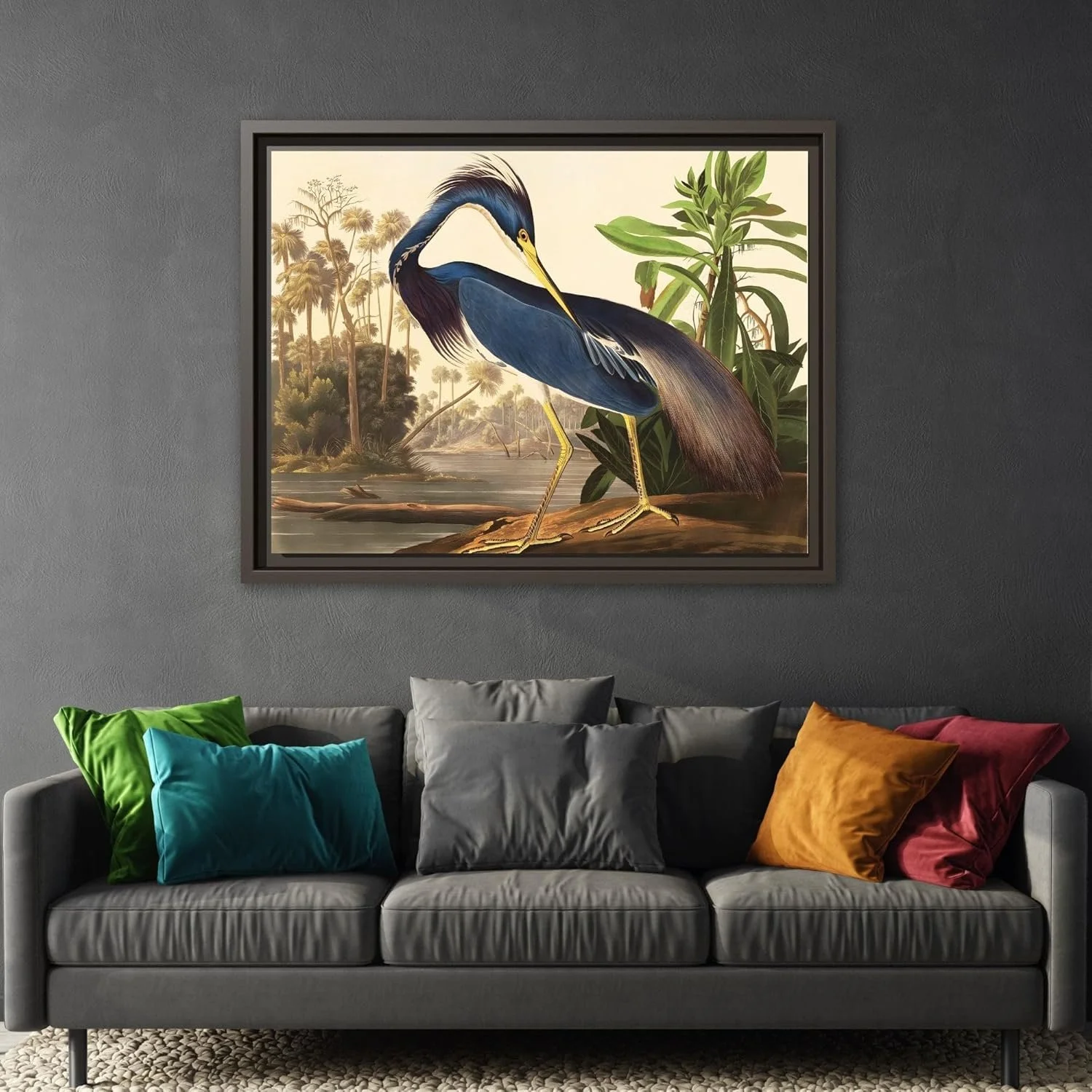 A framed artwork of a heron standing by water in a jungle setting hangs above a gray sofa decorated with six colorful pillows.