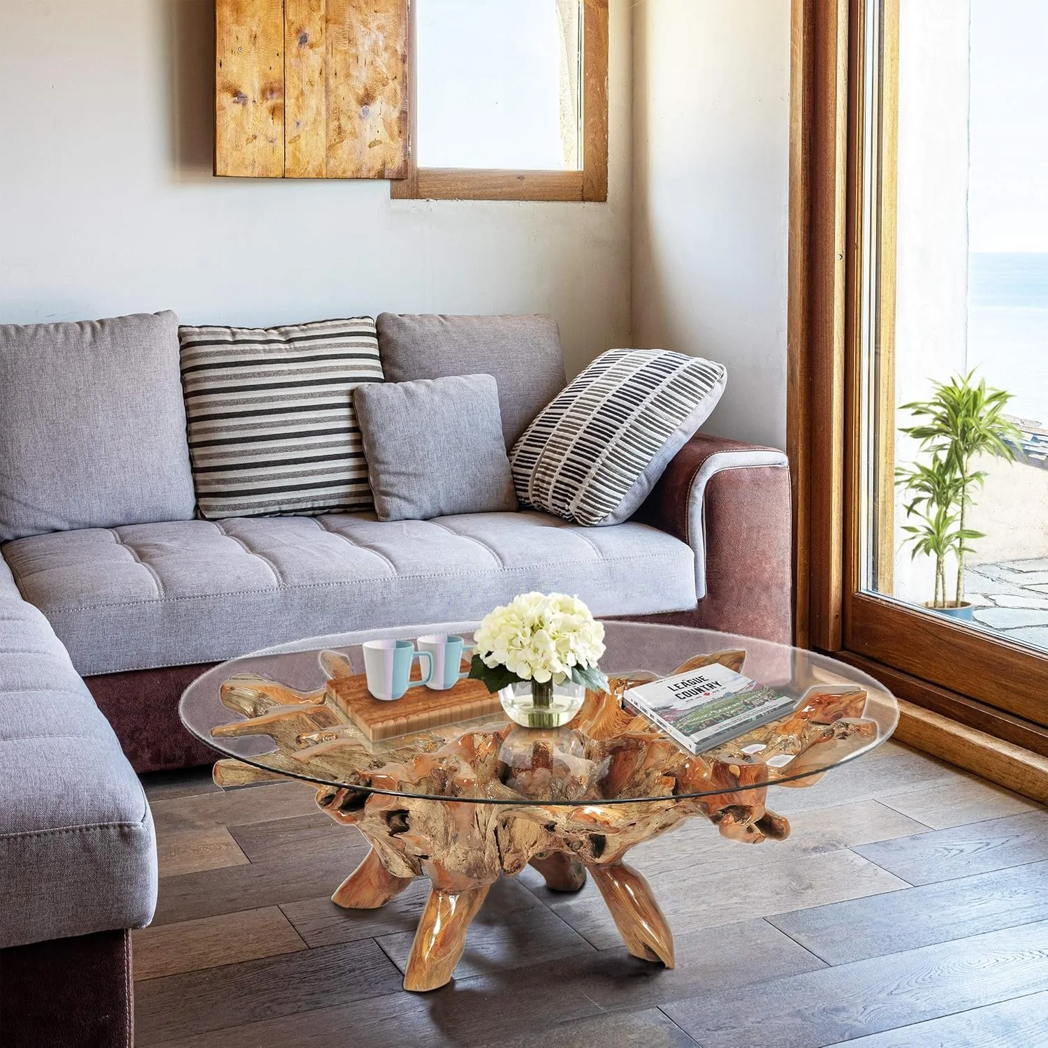 Living room with a light gray sofa, patterned pillows, a glass top coffee table with a wooden base, a white flower arrangement, and a large window with a view of the outside.