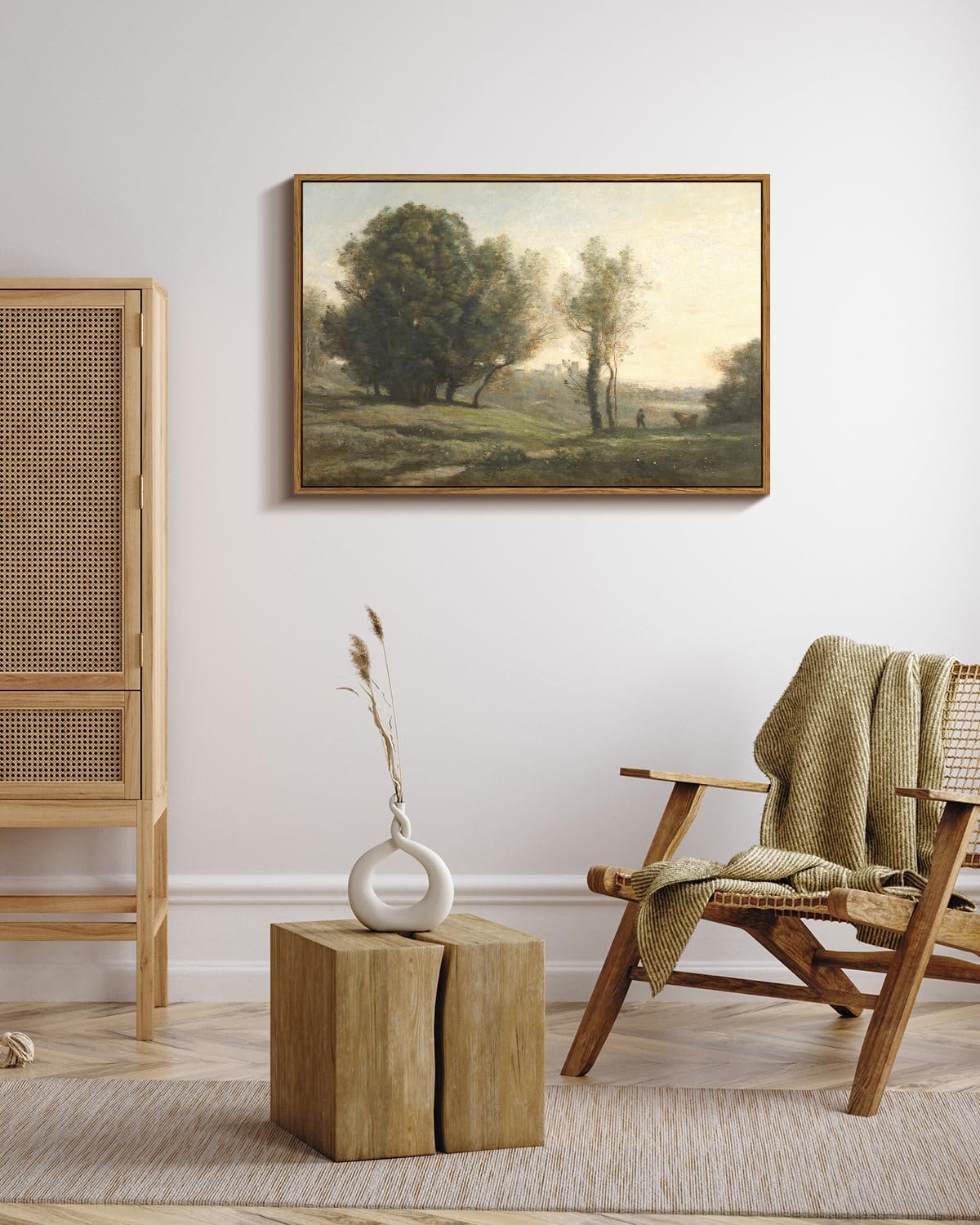 A living room with a framed landscape painting of trees on the white wall. A wooden cabinet with a woven front is on the left. A wooden table with a white decorative vase holding dried grass is in front. A wooden armchair with a beige woven blanket draped over it is on the right.