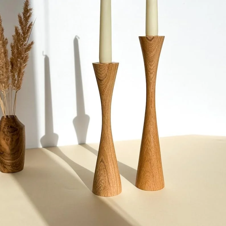 Two wooden candle holders with tapered candles on a light-colored surface, casting shadows on a white background, with a small wooden vase containing dried grasses on the side.
