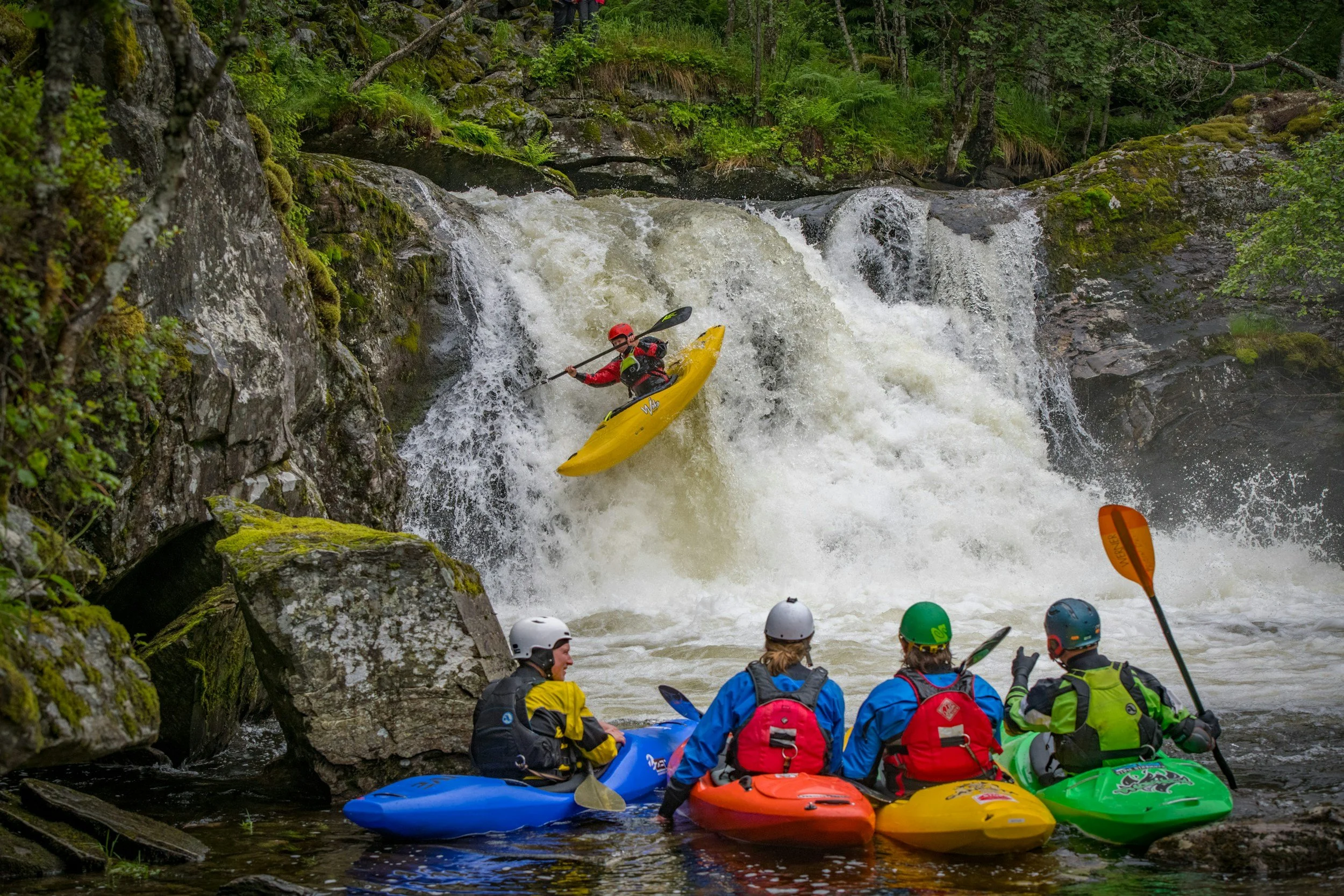 four white water kayakers watch as another kayaker rides down a small waterfall