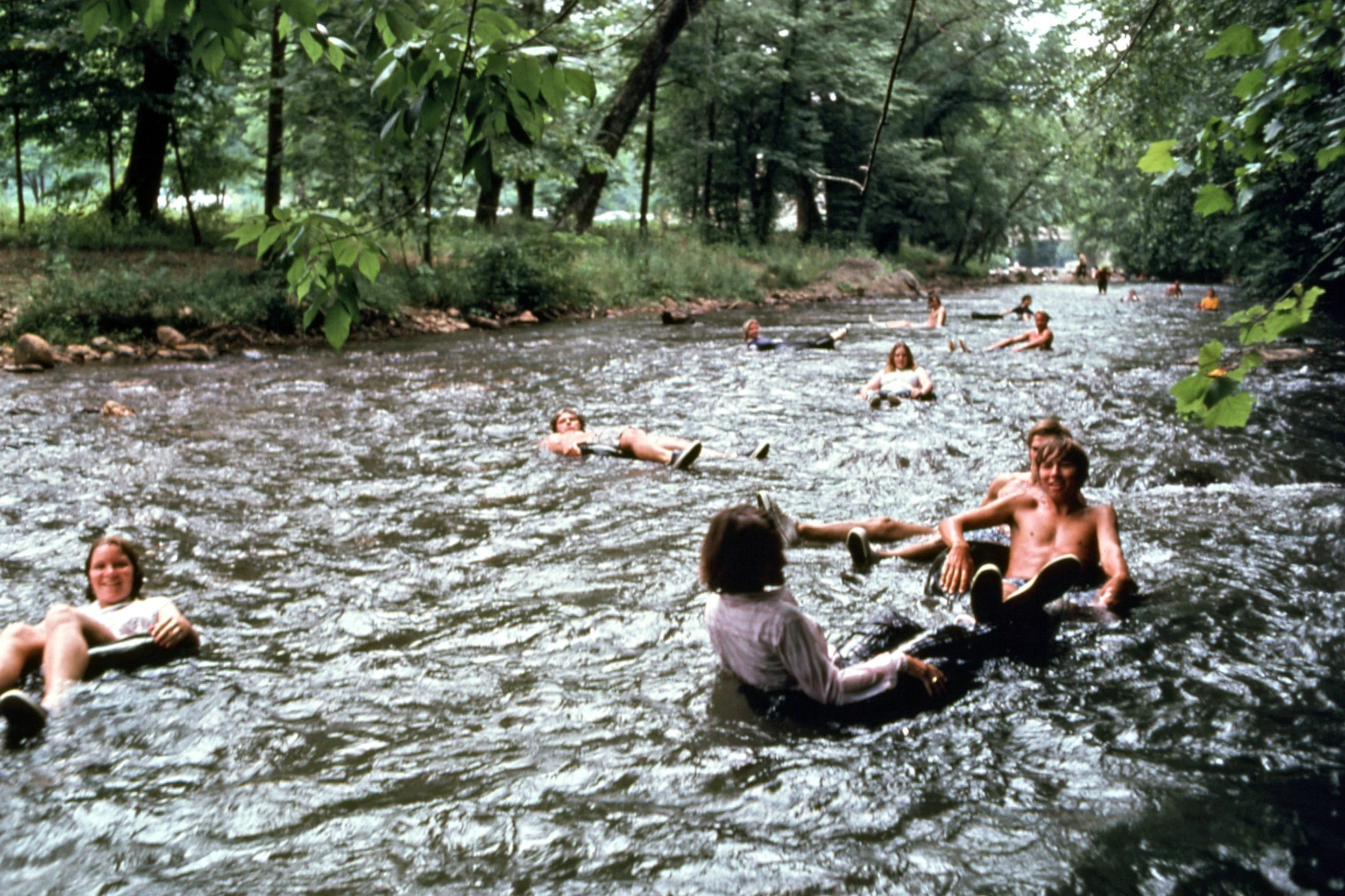people smiling while tubing comfortably down a river with green grass and trees on both sides