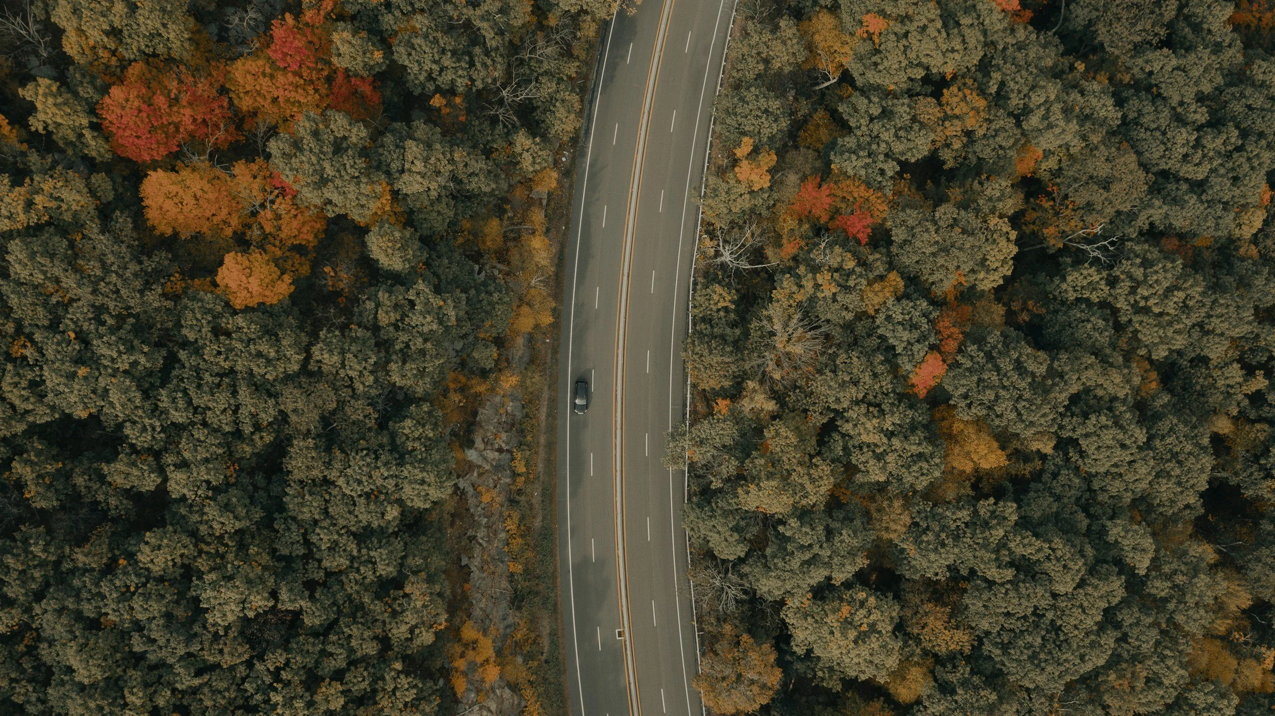 an aerial photo of  a car driving on an empty, winding highway surrounded by forest