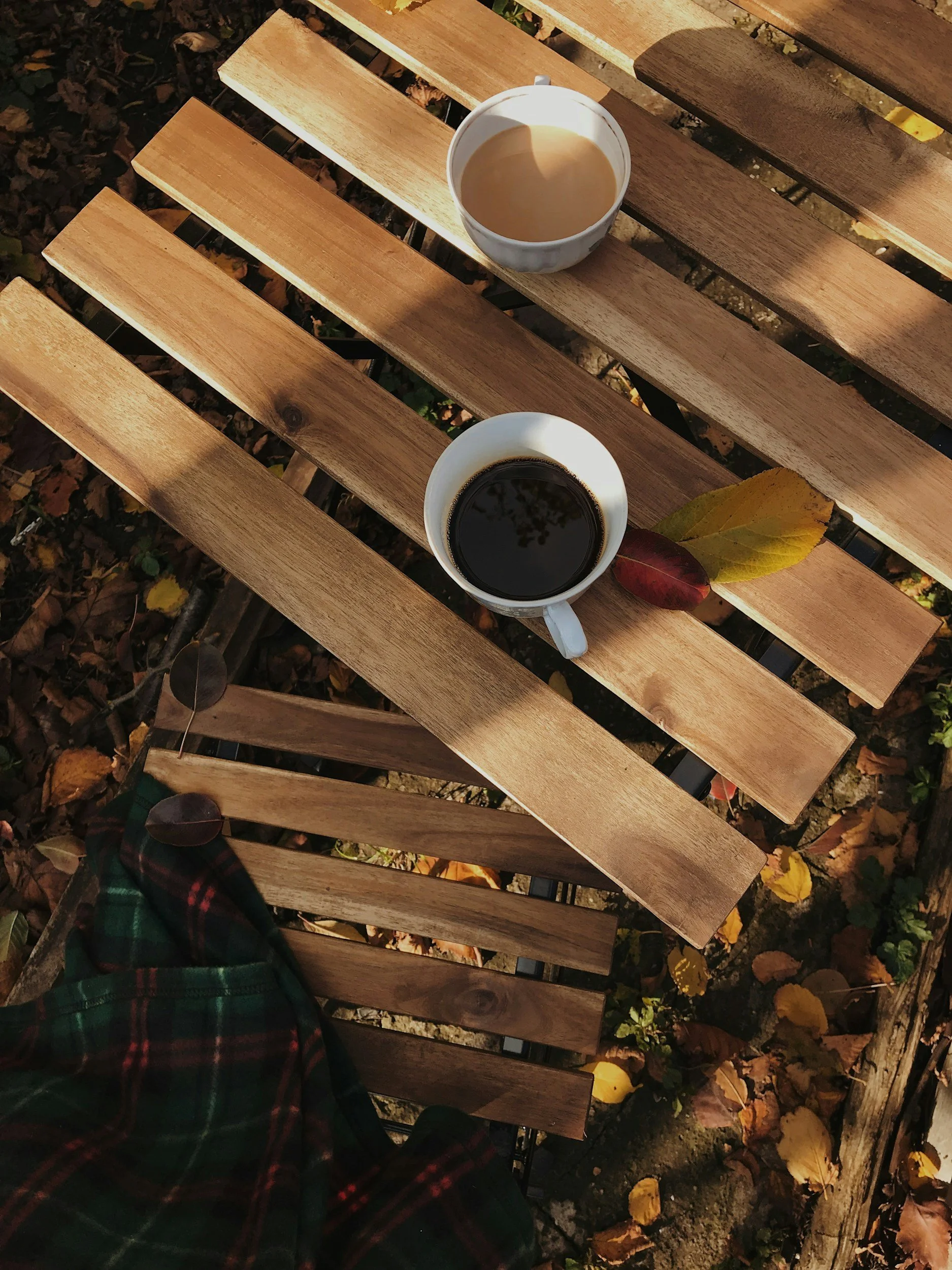 A cup of black coffee on a wooden table with fallen autumn leaves on the ground below