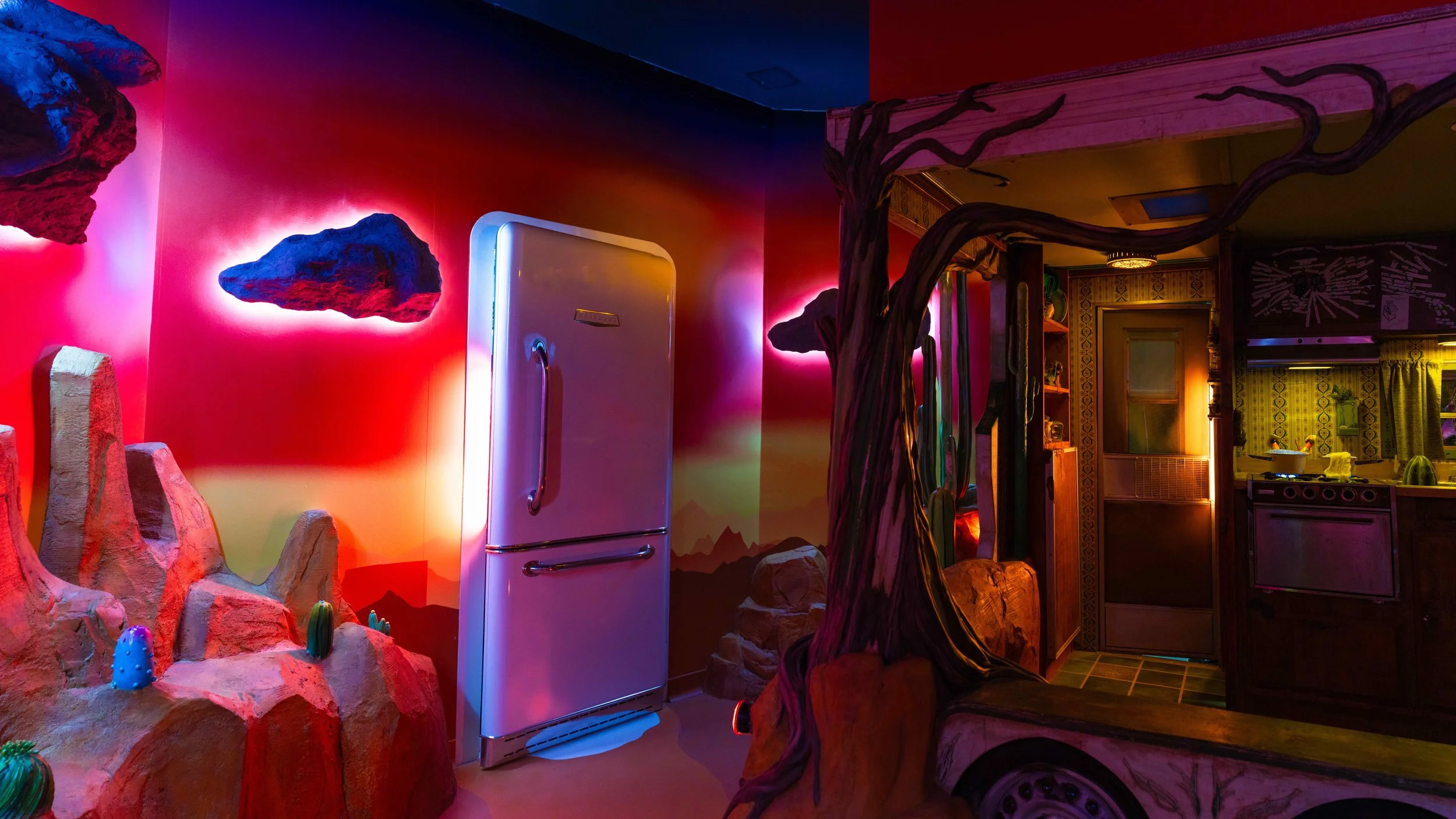 Press media image from Meow Wolf of an interior room that has an abstract desert setting mixed with a glowing fridge and glowing clouds