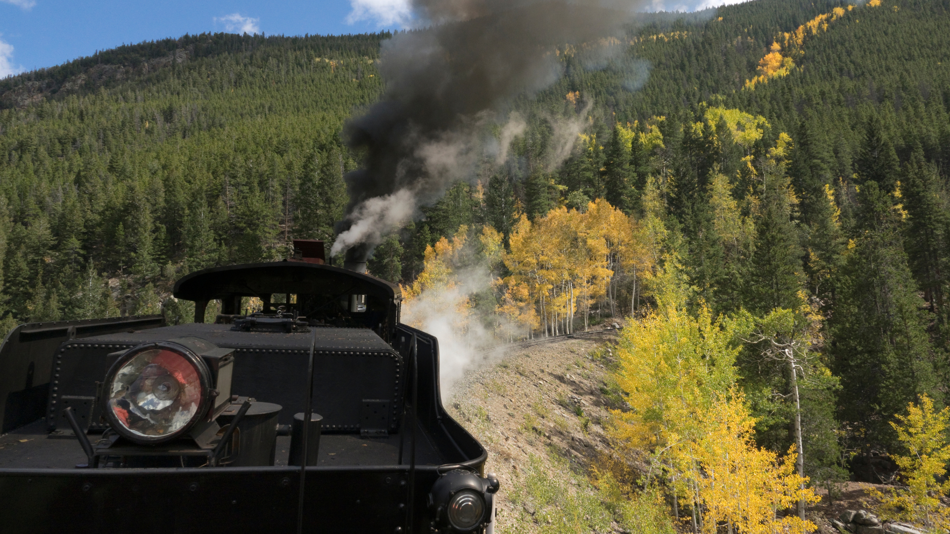 Puffing out black smoke, a steam locomotive pulls train cars over the steep grade on the Georgetown Loop Railroad in the Colorado Rocky Mountains